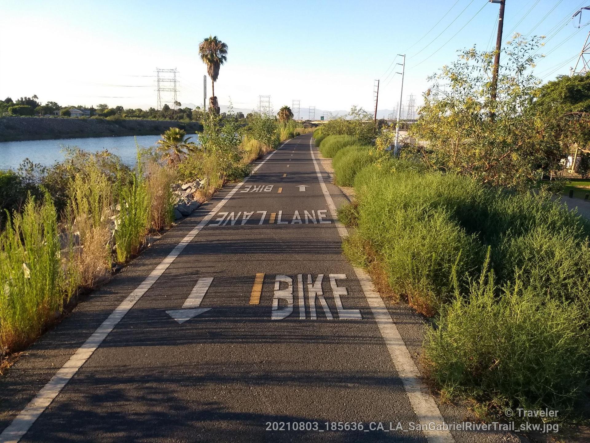 Paved bike path with "BIKE LANE" markings, green foliage, and river.