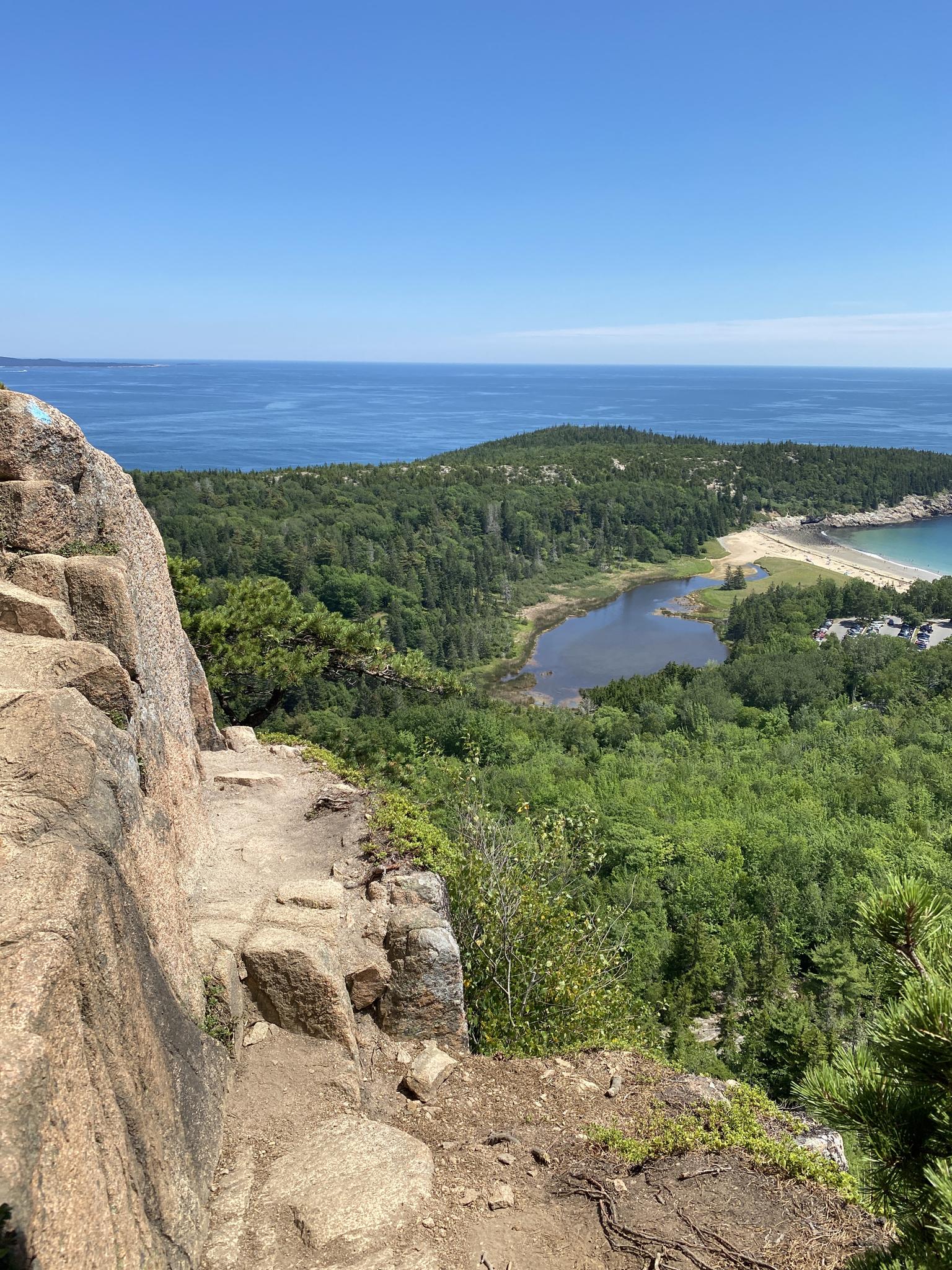 Sunny coastal view from rocky cliff, showing forest, lake, beach, and ocean.