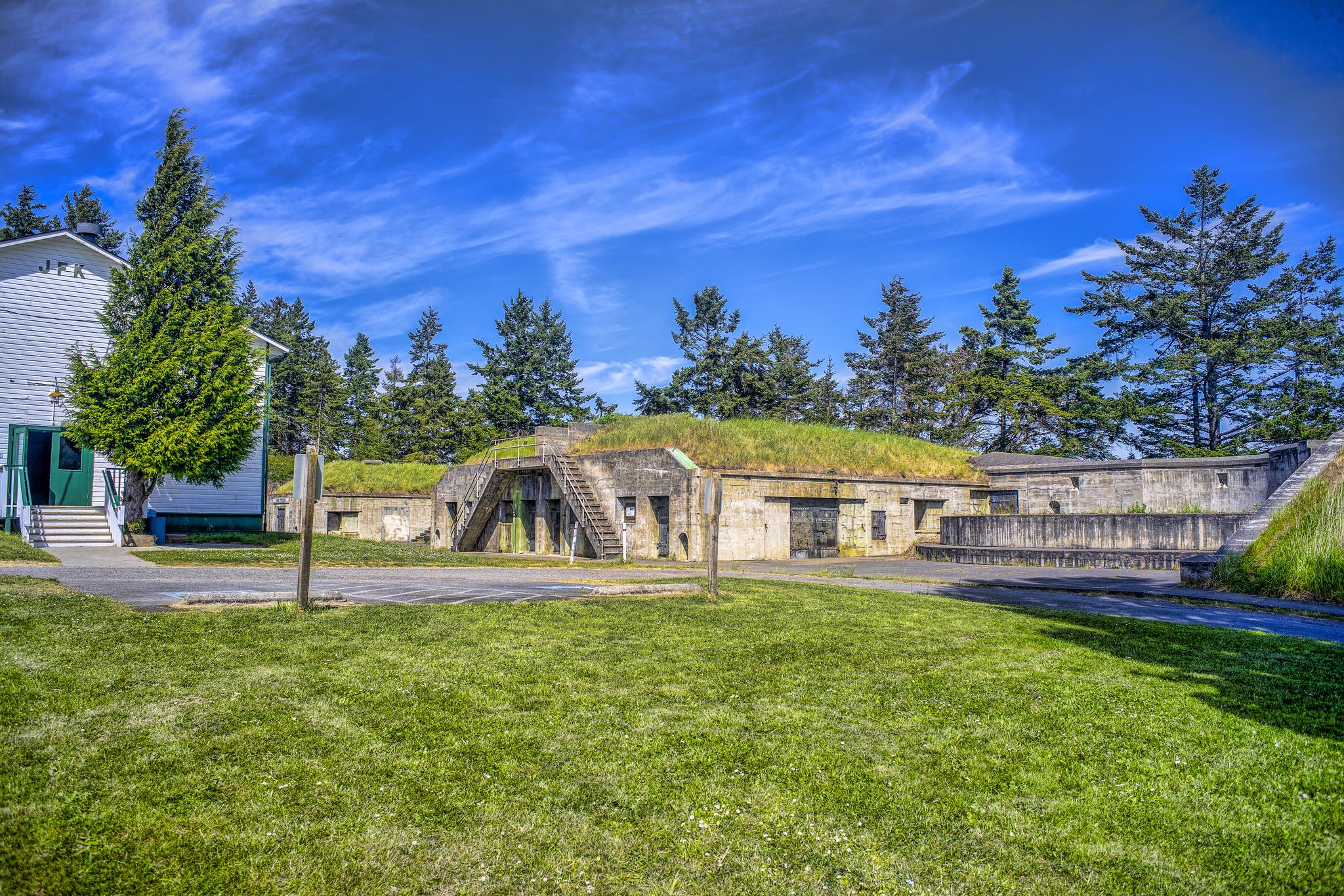 Old concrete bunkers with grass roofs and a white building under a bright blue sky.