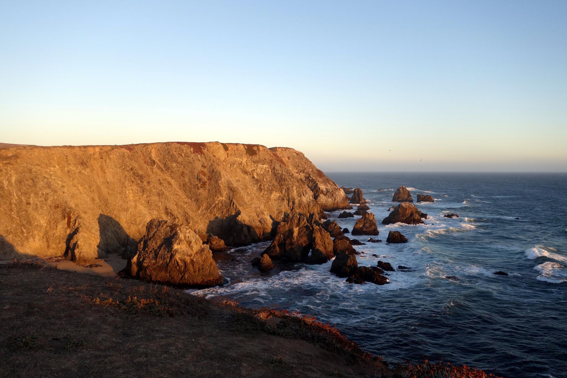 Rugged sea cliffs and sea stacks at sunset, with ocean waves.