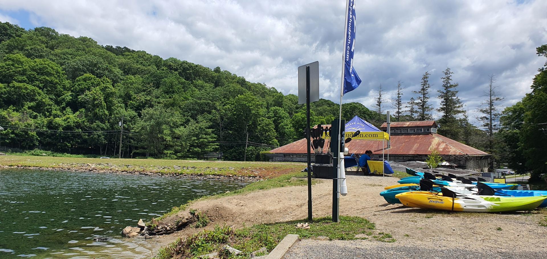 Colorful kayaks on a lakeshore next to a building, with forested hills.