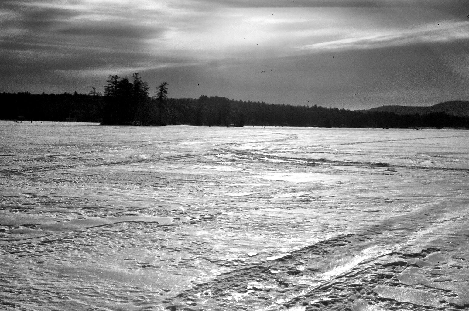 Black and white image of a vast snowy field with tracks, dark forest.