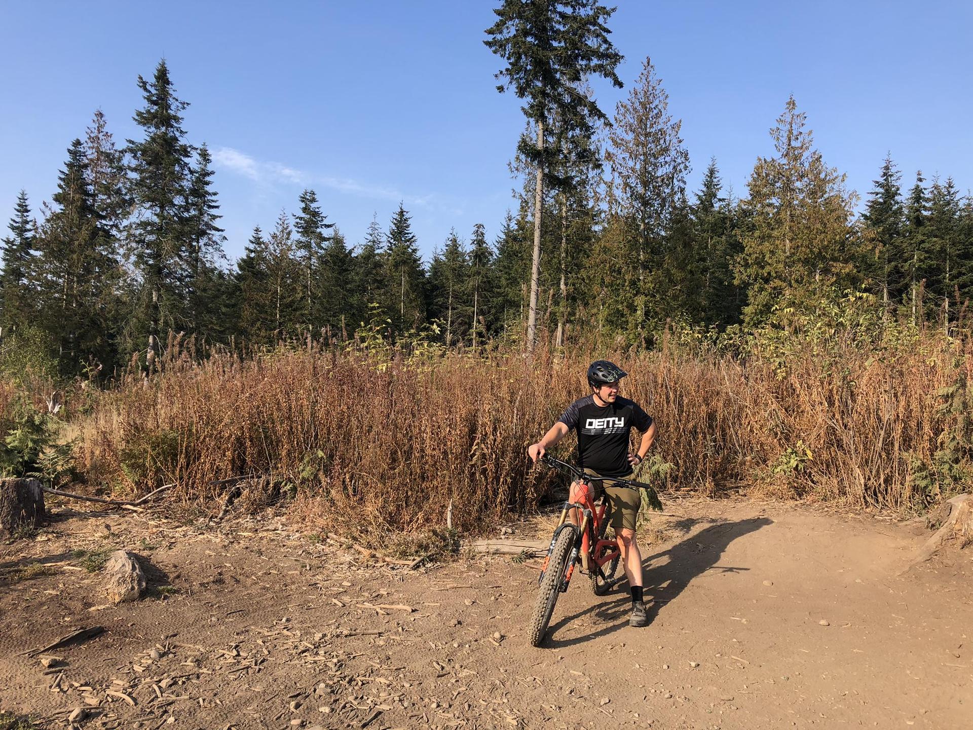 Mountain biker standing on a sunny dirt trail surrounded by trees and dry grass.