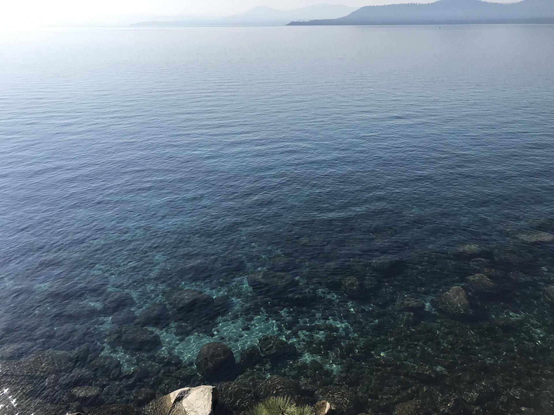 Clear blue lake, submerged rocks, hazy distant mountains, overcast lighting.