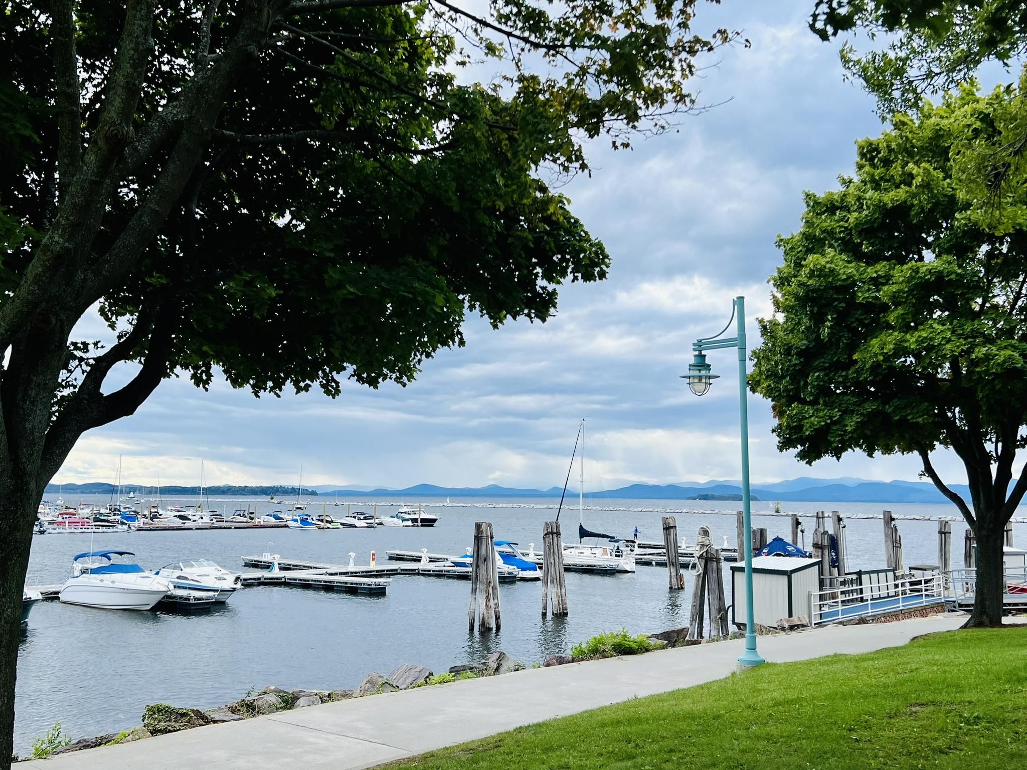 Boats docked in a marina, framed by green trees, with distant mountains and cloudy sky. - Cycle the City and Intervale Trail