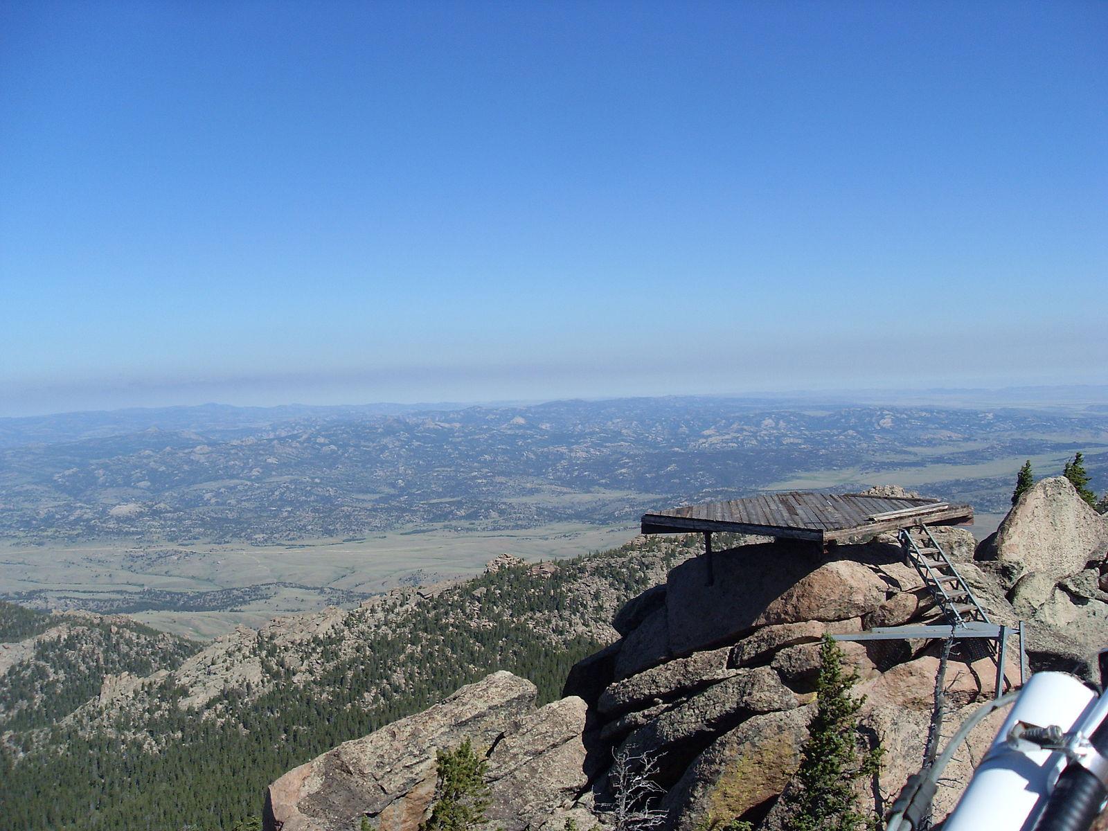 Wooden platform on rocky peak overlooking vast hazy mountain landscape under clear sky.