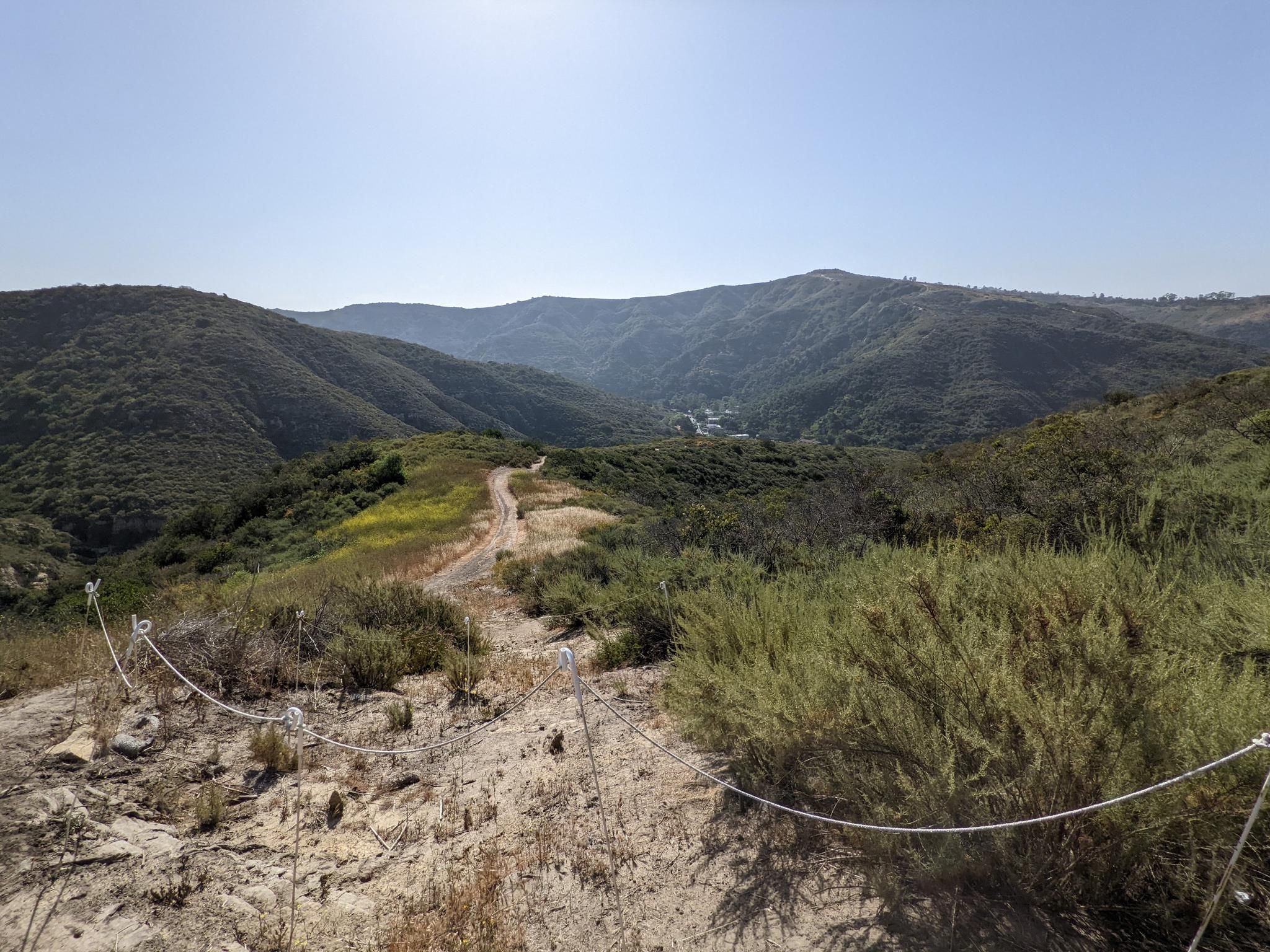 A dirt path descends into a green valley under a bright, sunny sky. - Laguna Coast Wilderness Park – Laguna Ridge Trail loop from Laguna Coast Wilderness Park