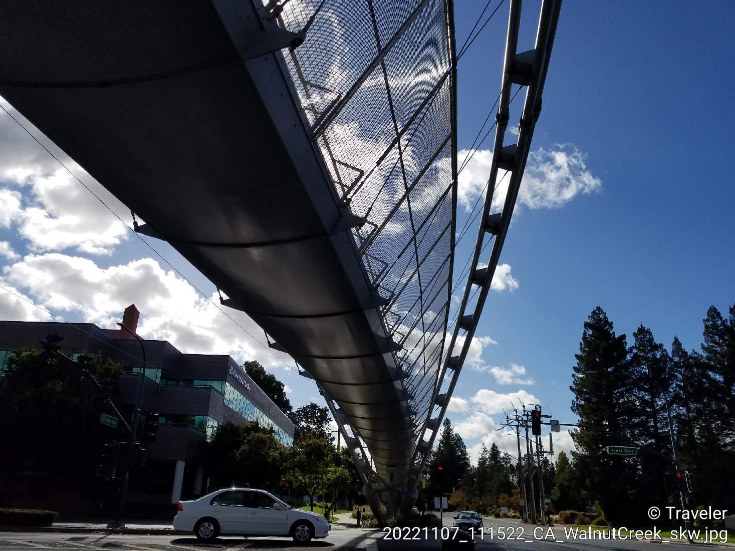 Modern pedestrian bridge over a road with cars, under a bright, cloudy sky.