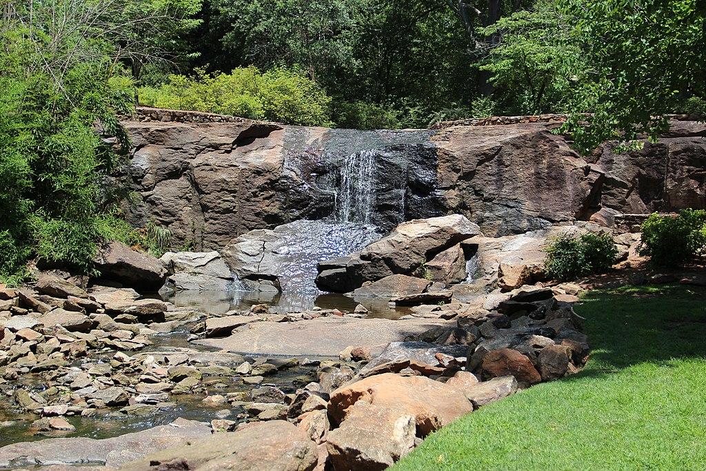 Sunny waterfall cascading over dark rocks into a shallow stream with green foliage.