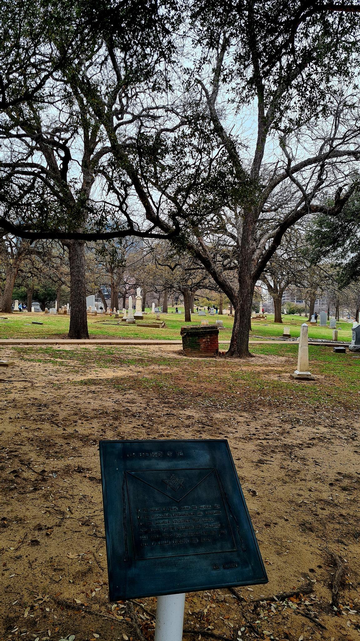 An information plaque in the foreground of a tree-filled, historic cemetery.