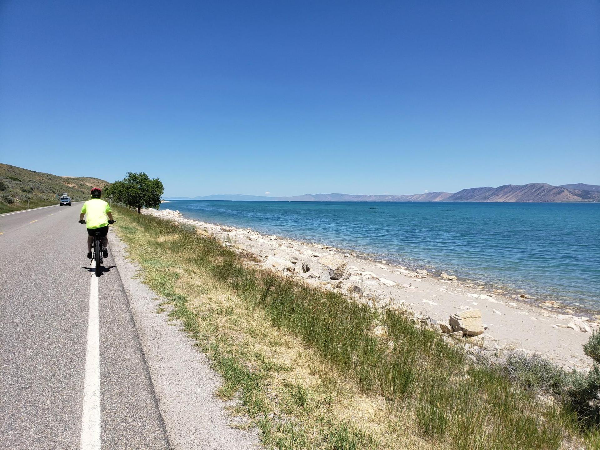 Cyclist on a sunny road next to a clear blue lake with distant hills.