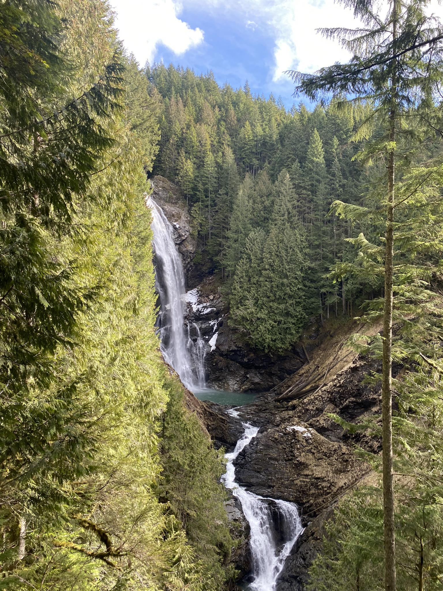A tall waterfall plunges into a deep, forested gorge under a bright sky.