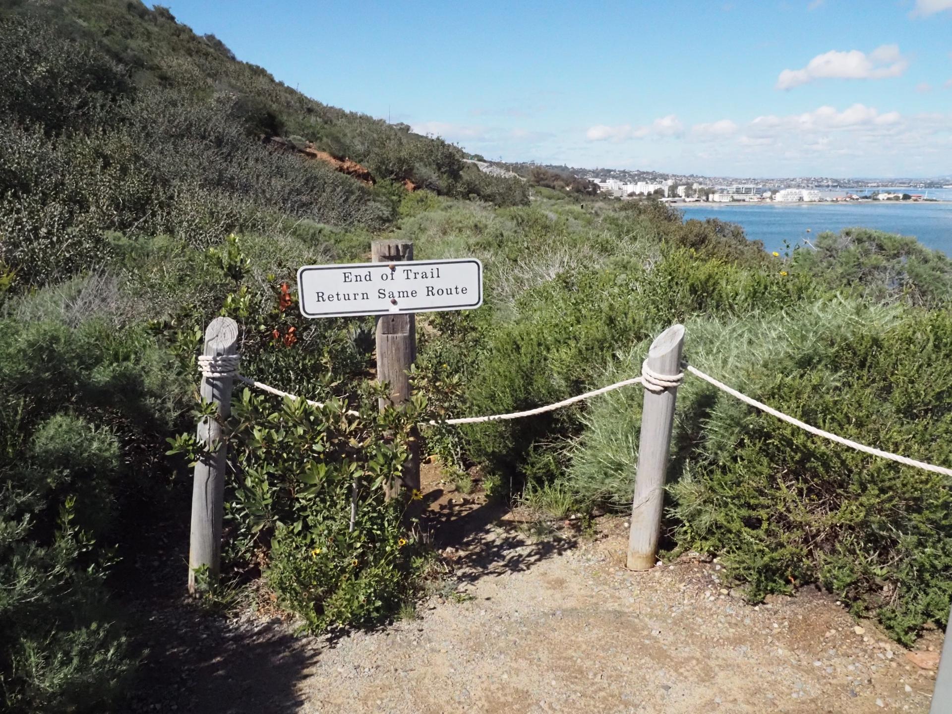 End of trail sign with rope fence on a bushy hillside overlooking water.