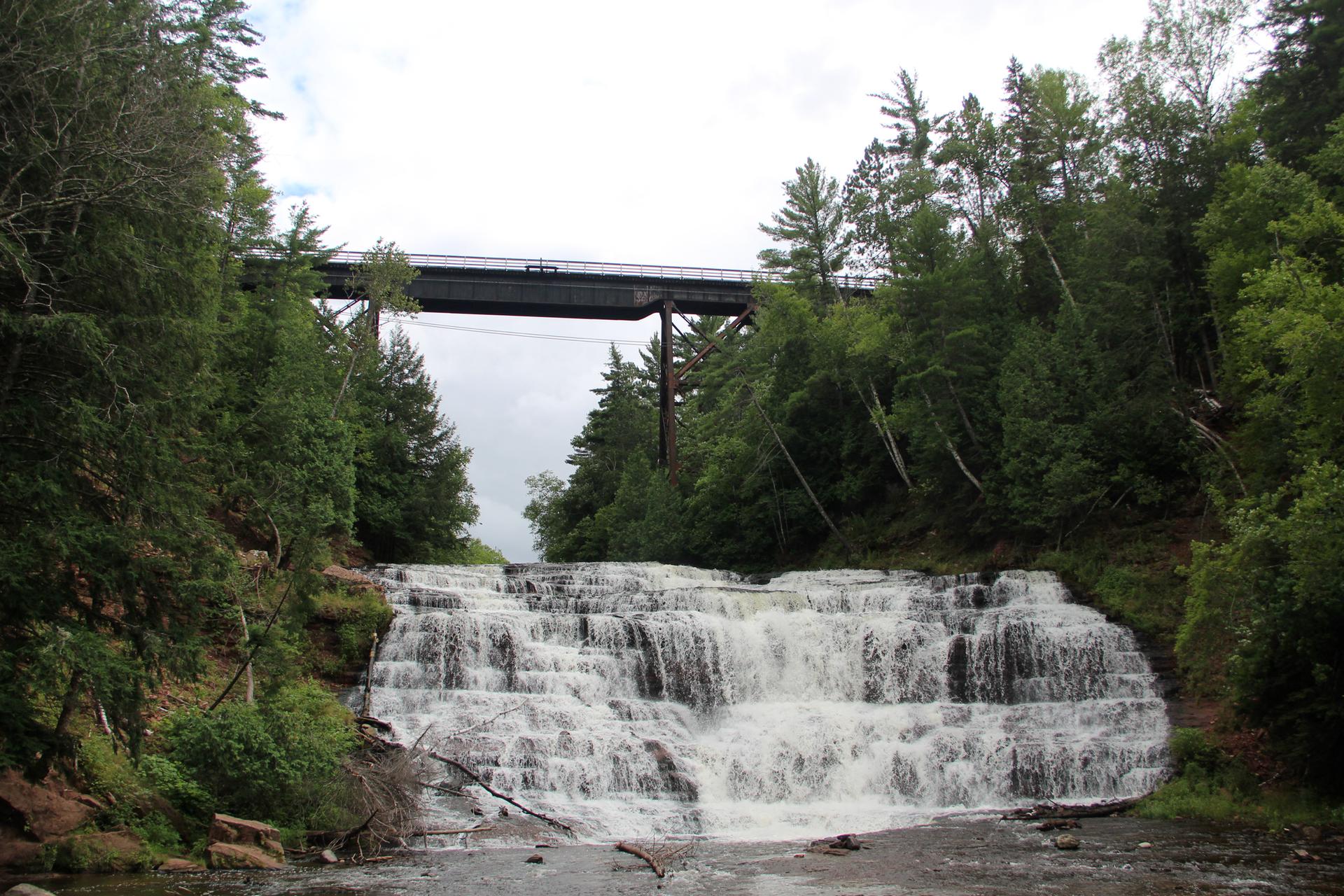 Wide waterfall cascading under a bridge, surrounded by lush green forest.