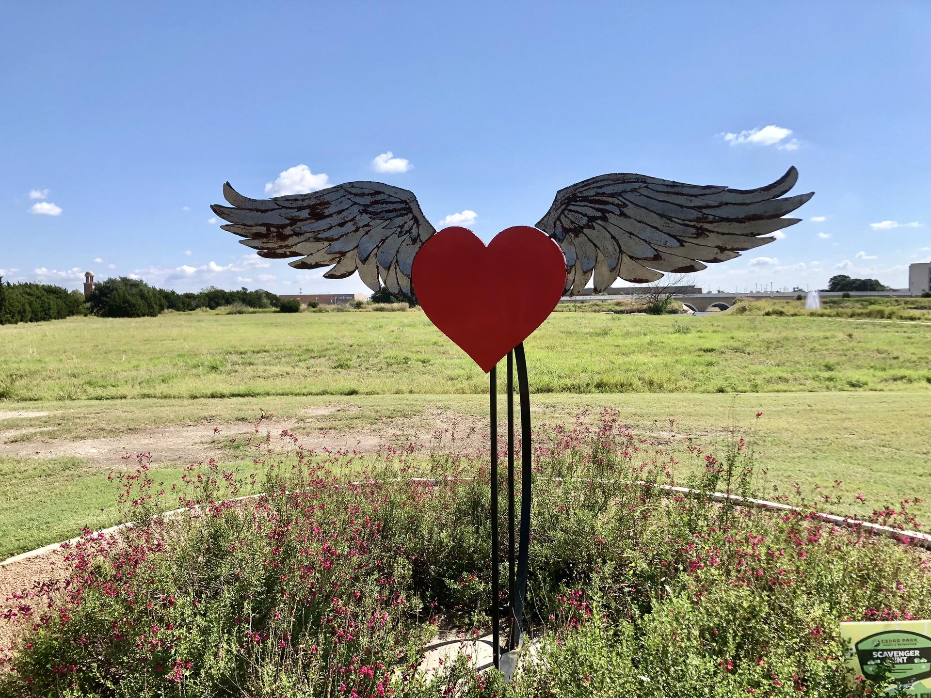 A red heart sculpture with metal wings in a sunny garden with a grassy field.