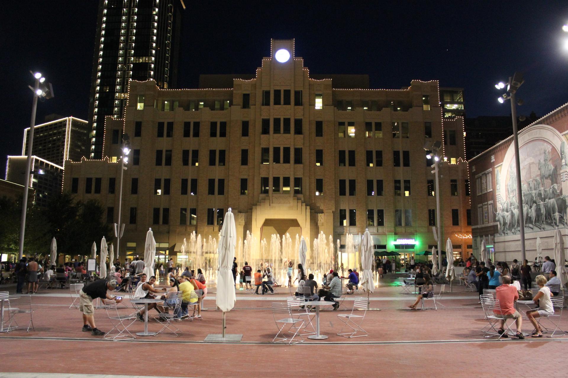 Crowded urban plaza at night with illuminated building, fountains, and people.