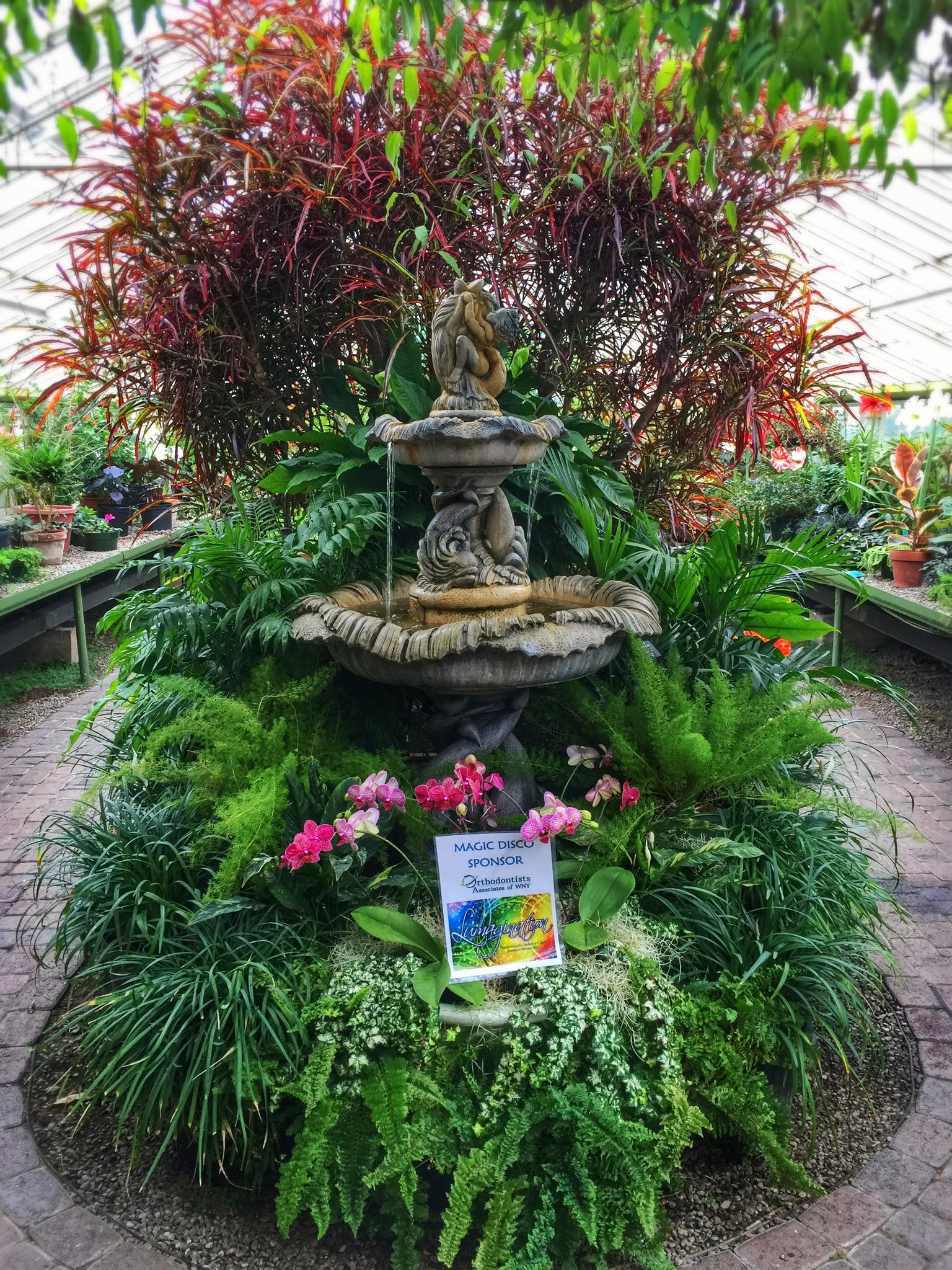 A multi-tiered stone fountain surrounded by lush green and red plants in a bright greenhouse.