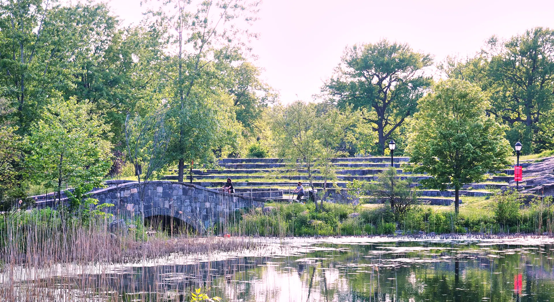 Sunny park scene with a stone bridge, pond, trees, and people relaxing.