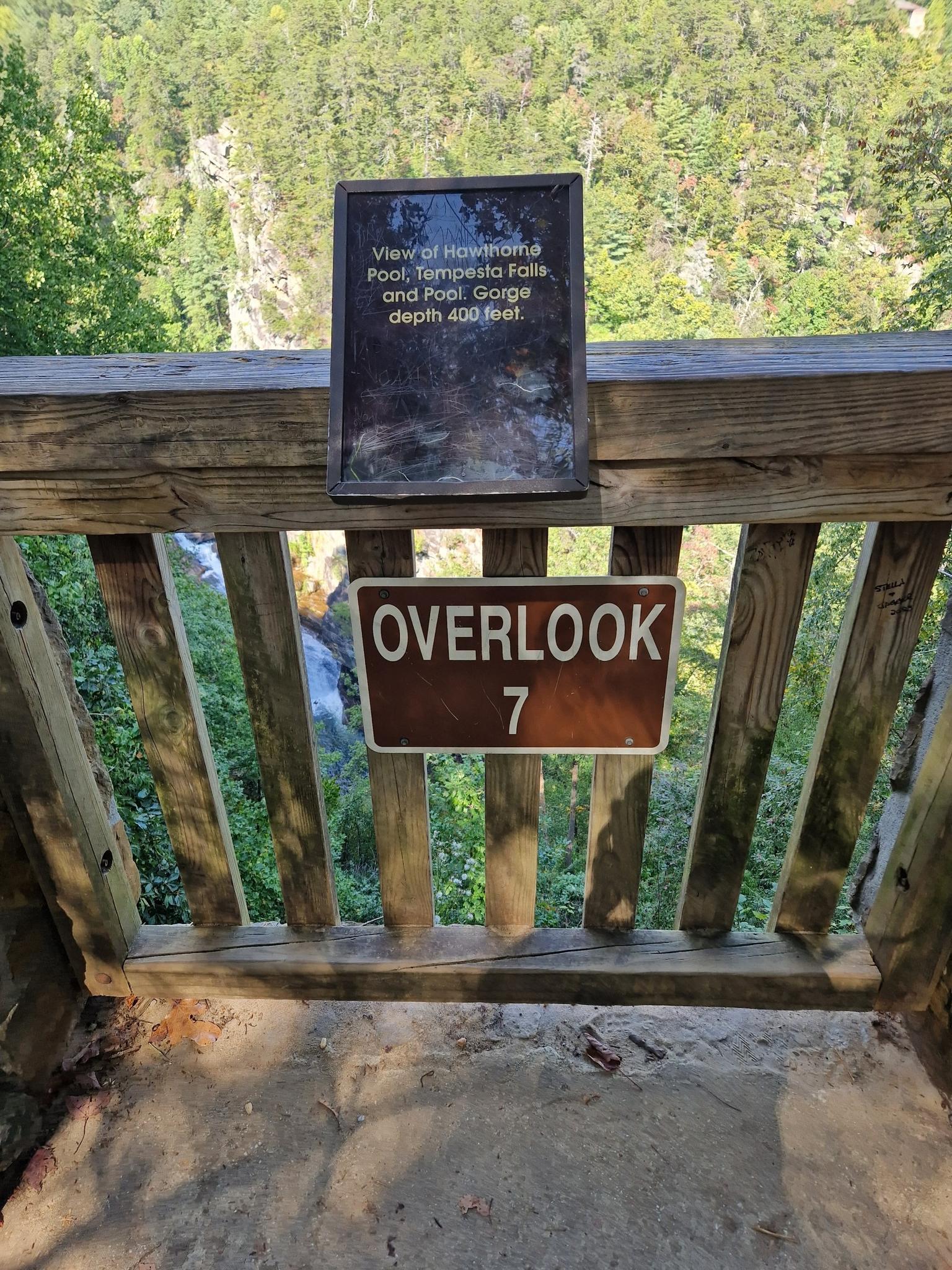 Wooden overlook railing with two signs, overlooking a green valley in daylight.