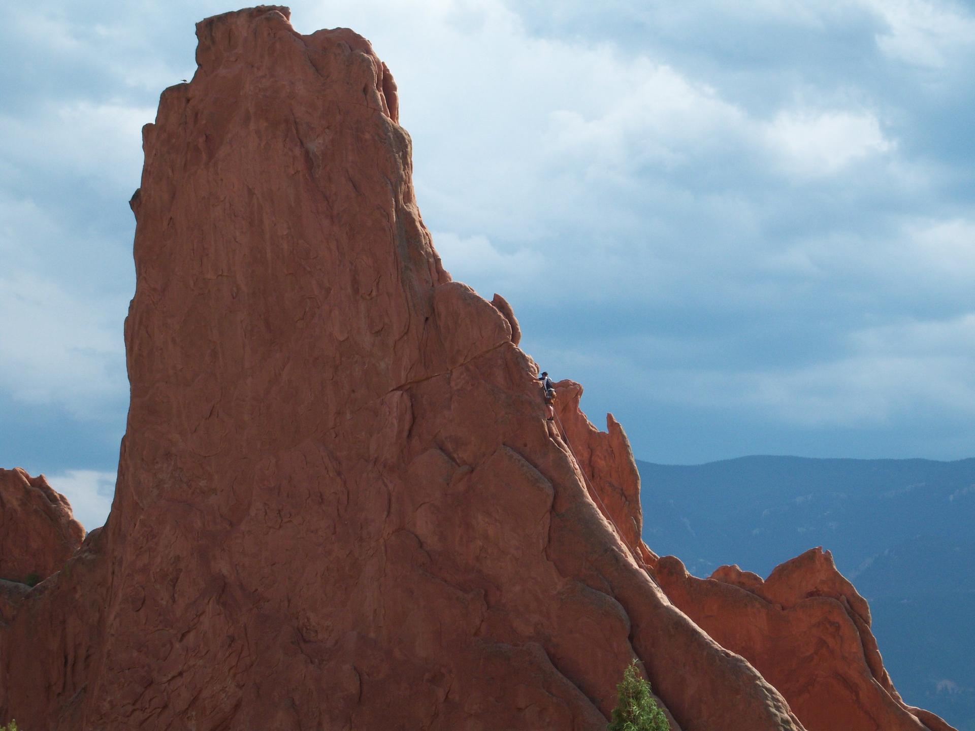 A person is rock climbing on a large red rock formation under a cloudy sky.