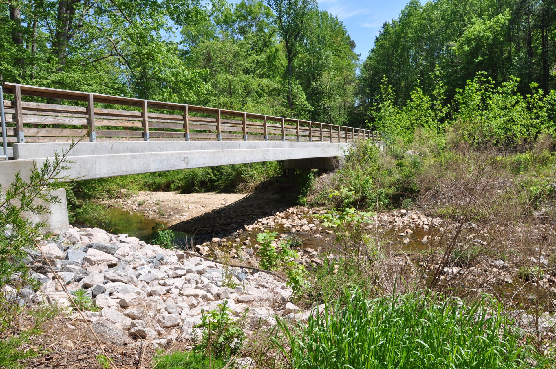 A concrete bridge with wooden railings crosses a rocky creek under bright daylight.