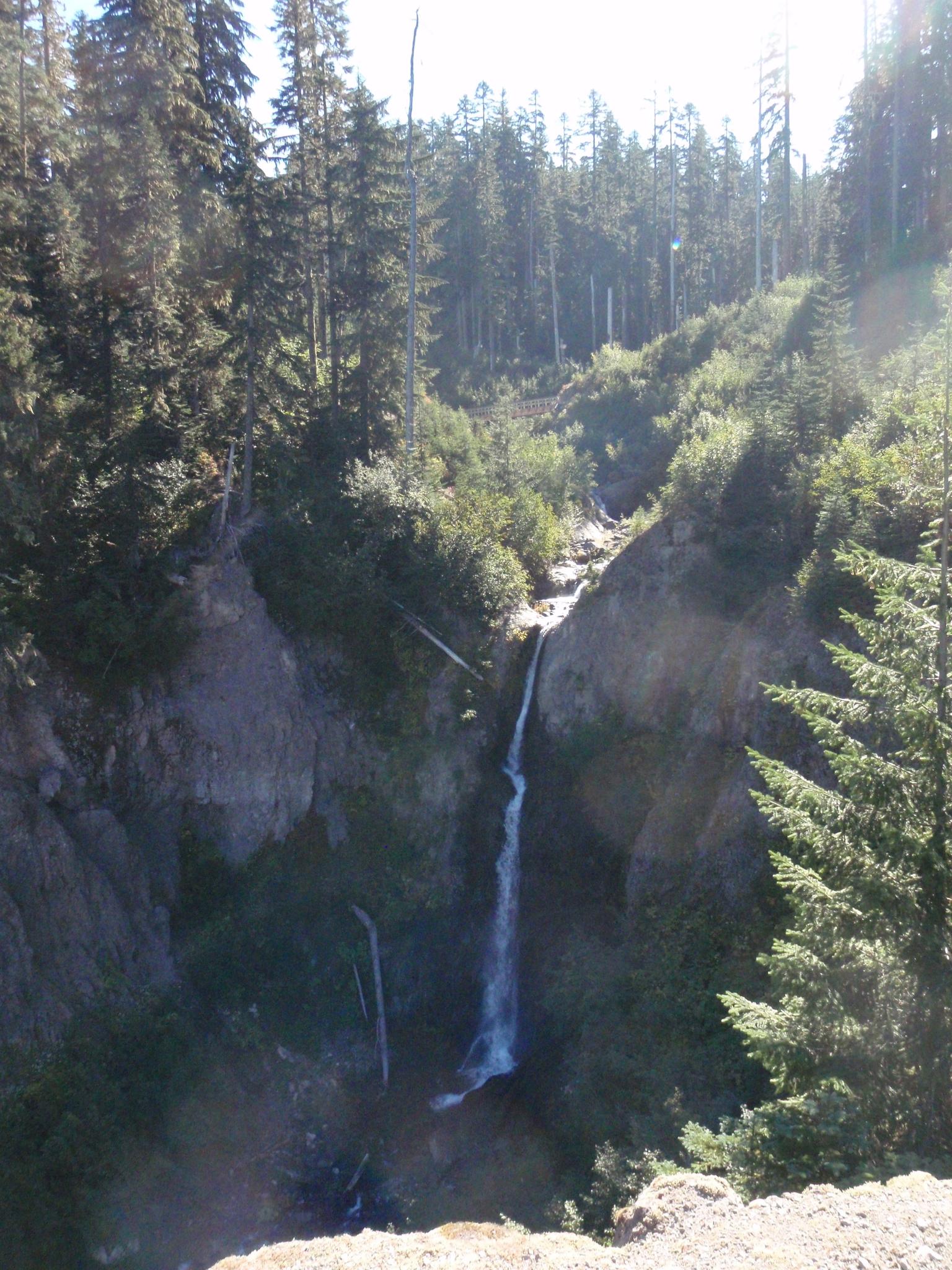 Tall waterfall plunges down a rocky, forested canyon on a sunny day.