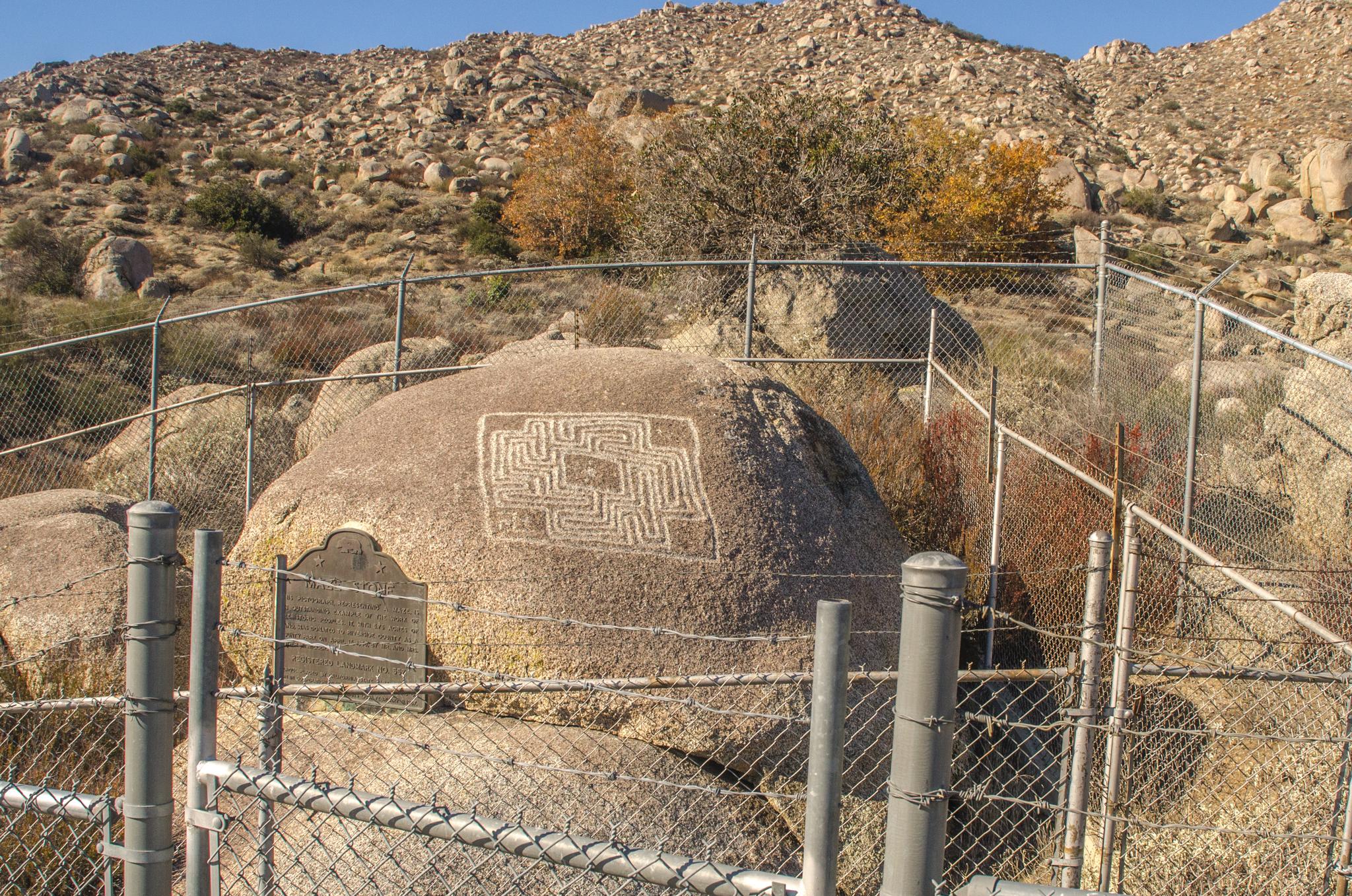 Ancient rock carvings and a plaque enclosed by a fence on a sunny, rocky hillside.
