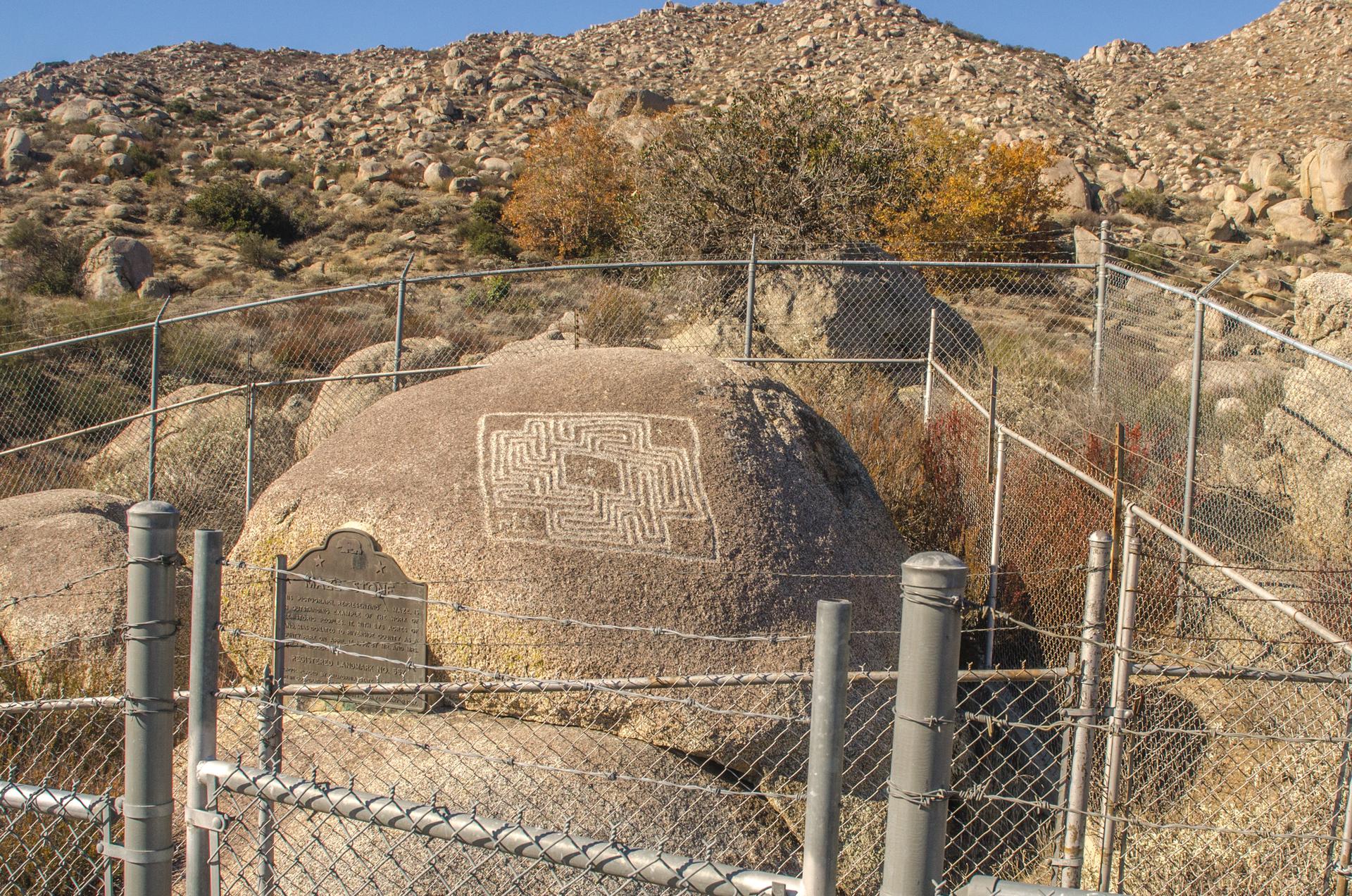 Ancient rock carvings and a plaque enclosed by a fence on a sunny, rocky hillside.
