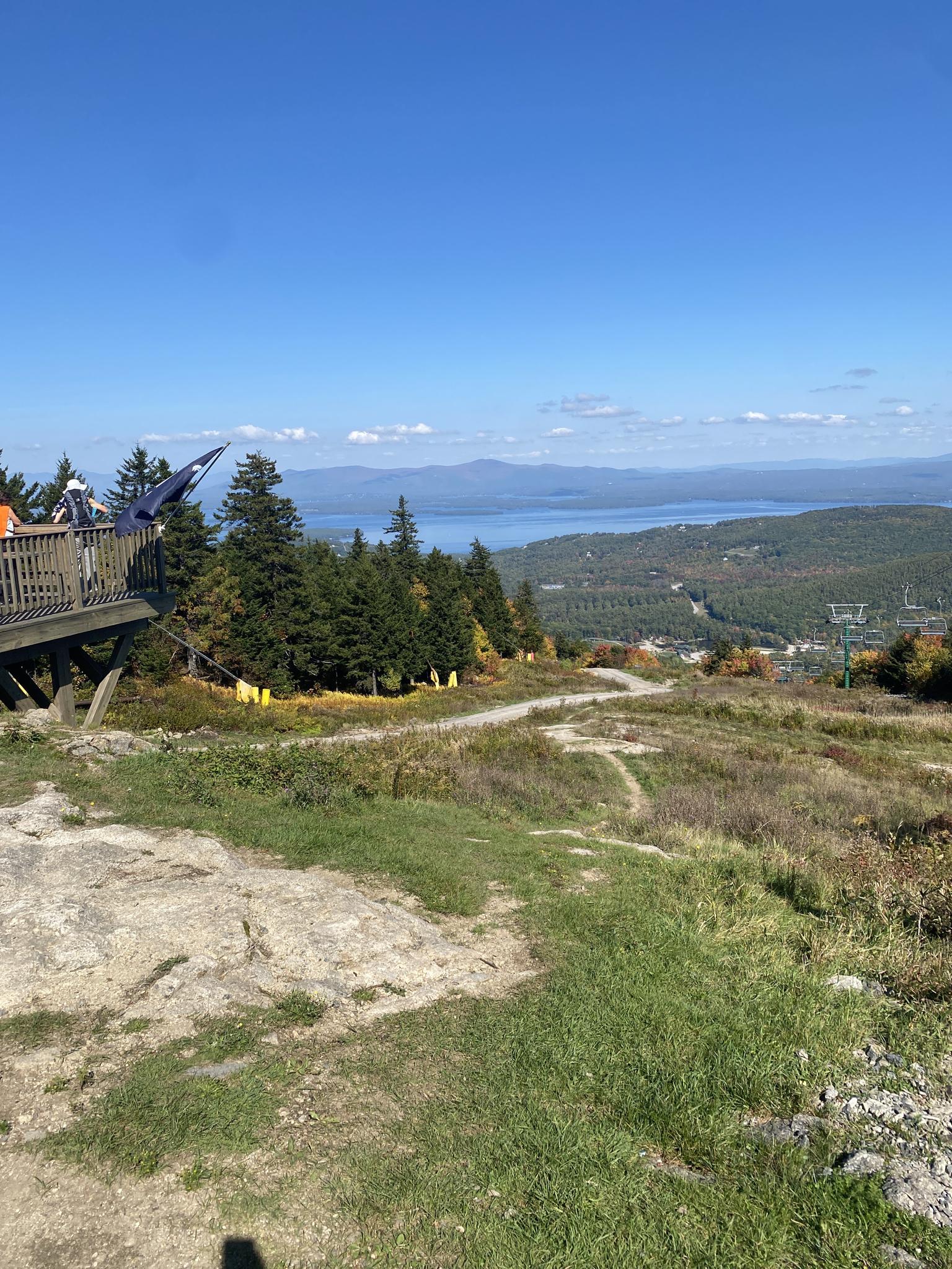 People on a wooden platform overlooking a distant lake and forested mountains.