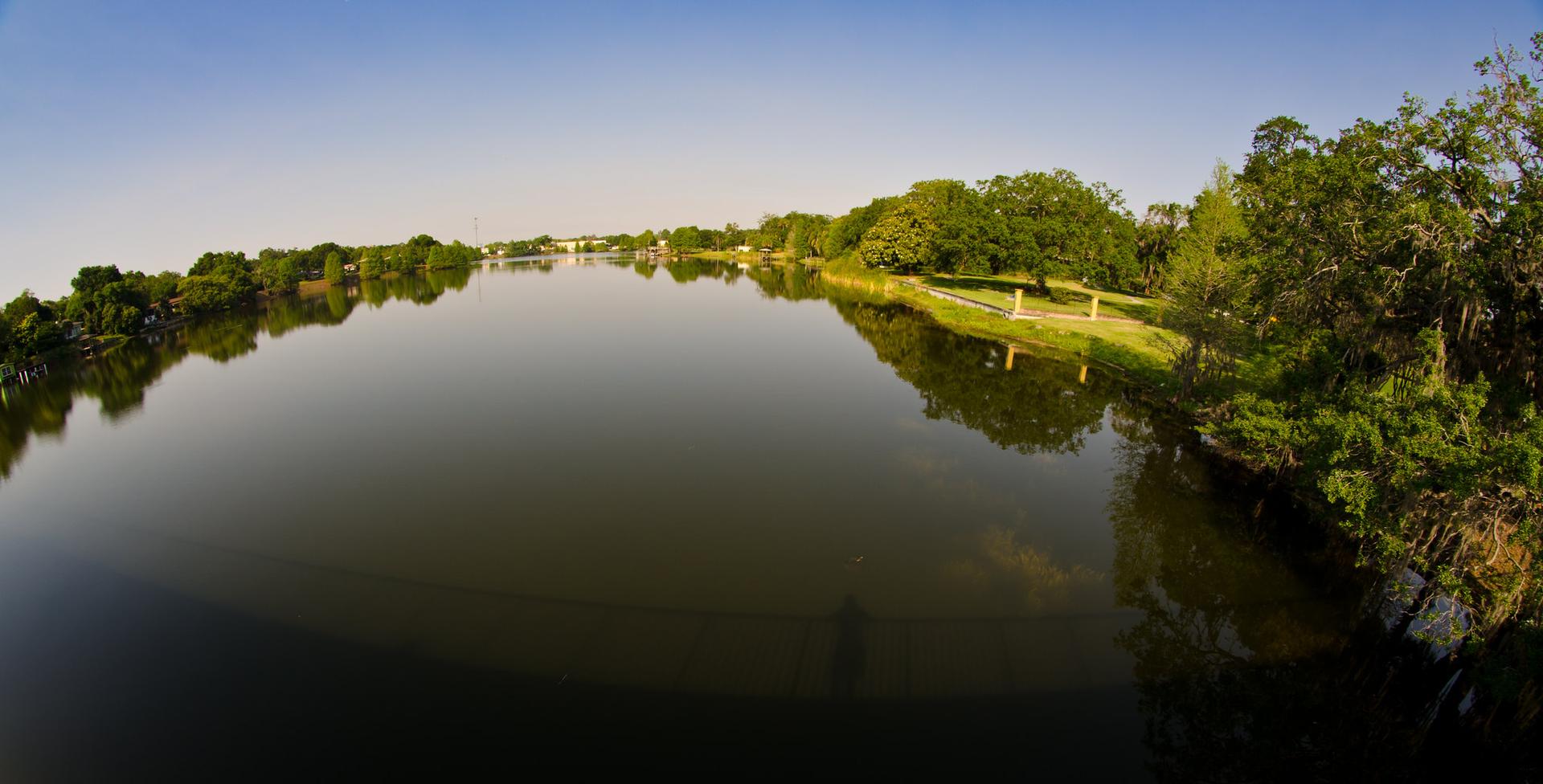 A wide, calm river reflecting green trees under a bright sky.