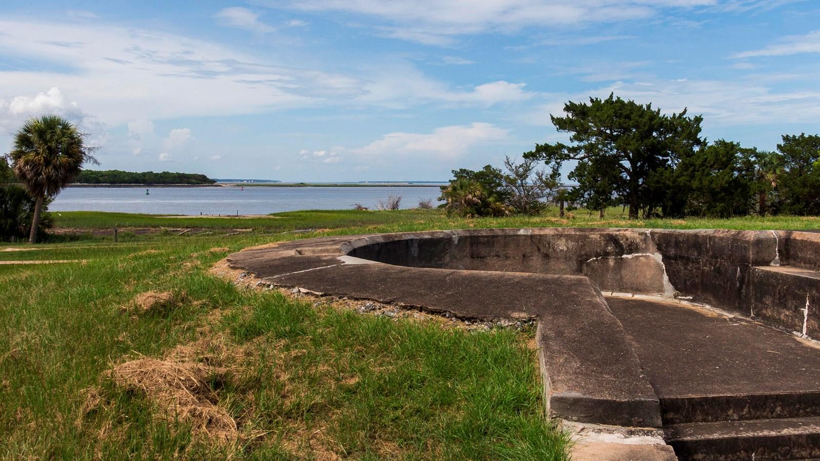 Concrete structure on grass overlooking water and distant land under a cloudy sky.