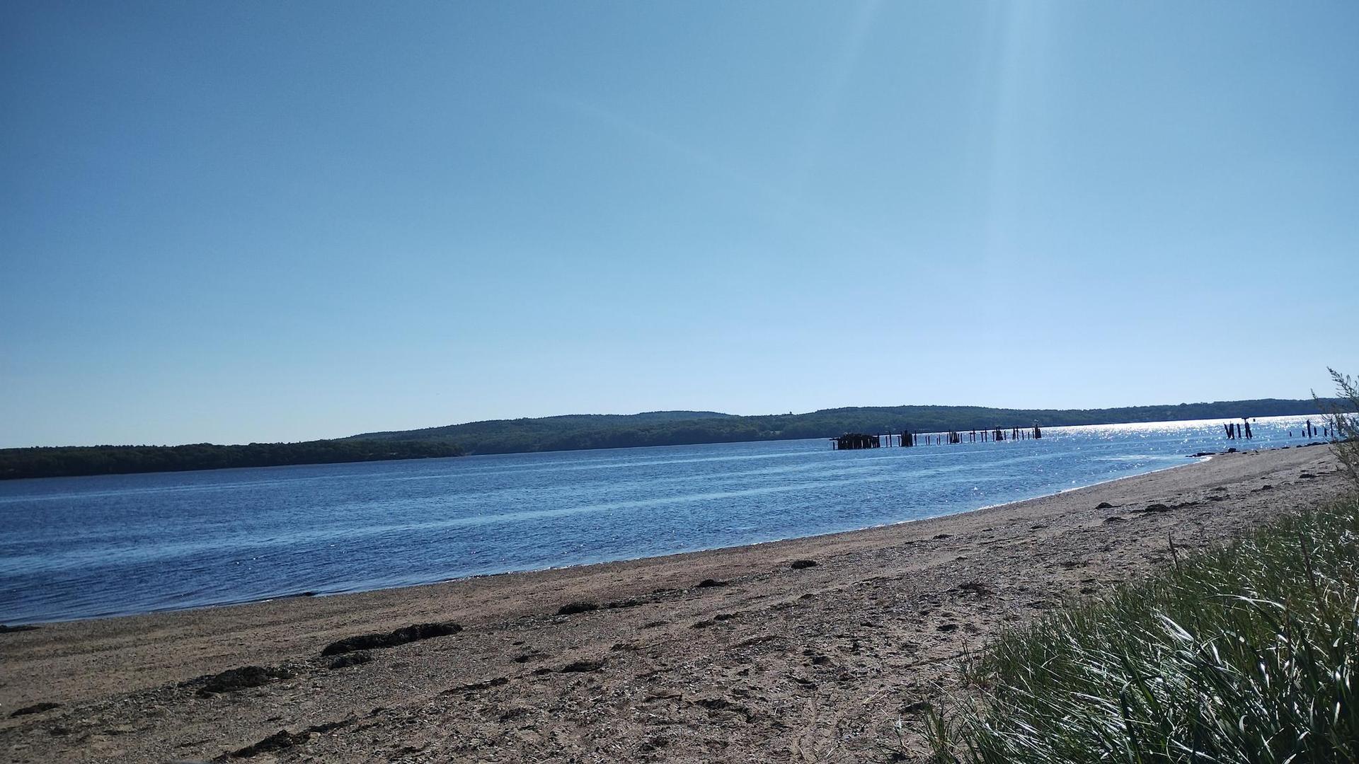 Wide sandy beach by calm blue water, distant forested hills, bright sky.