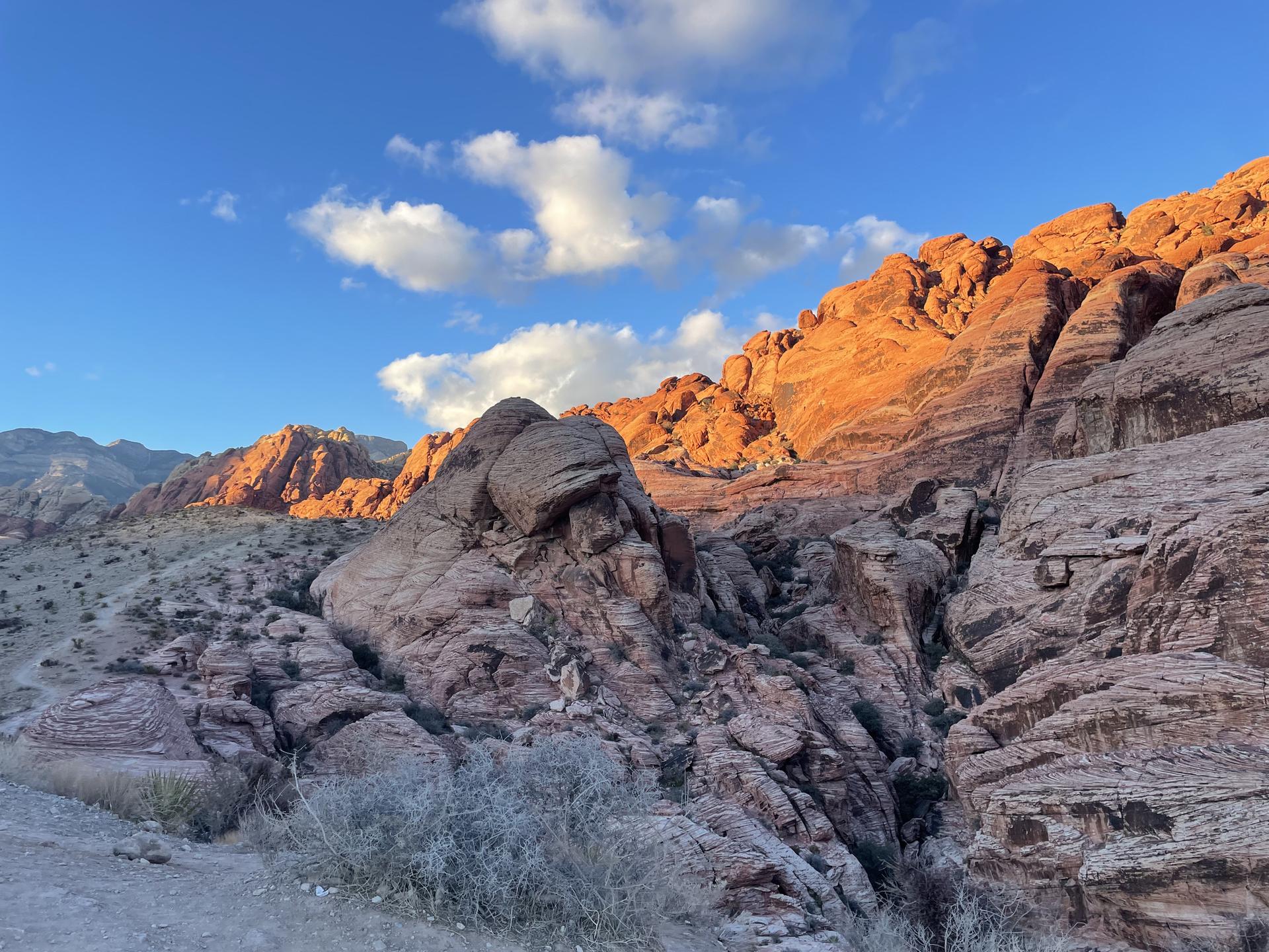 Golden light illuminates rugged red and tan desert mountains under a blue sky.