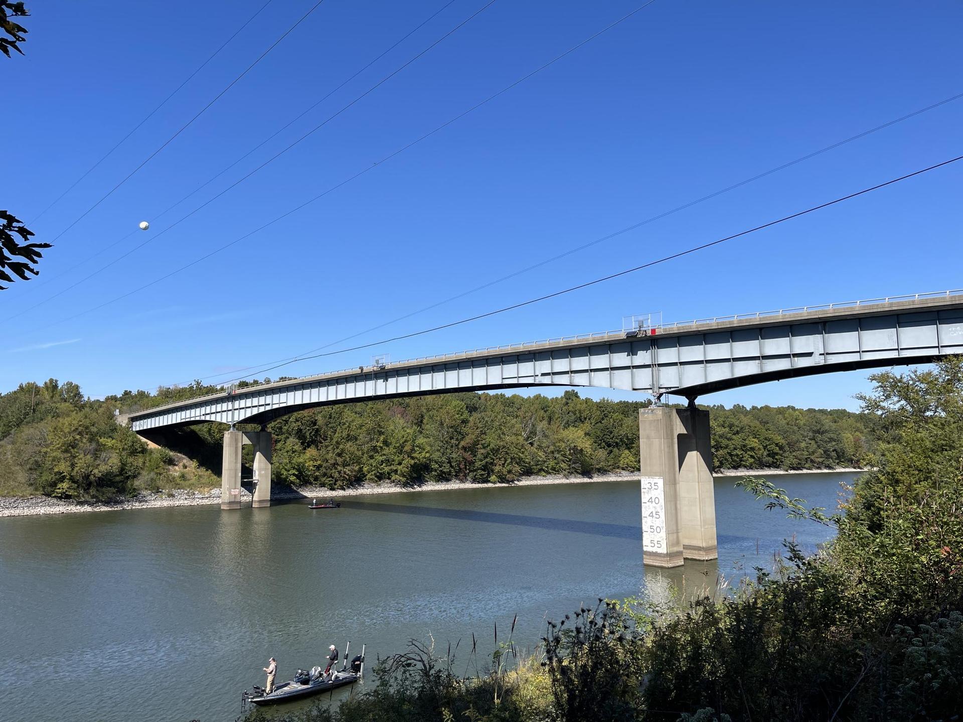A long bridge spans a river with forested banks under a clear sky. A boat with people is on the water.