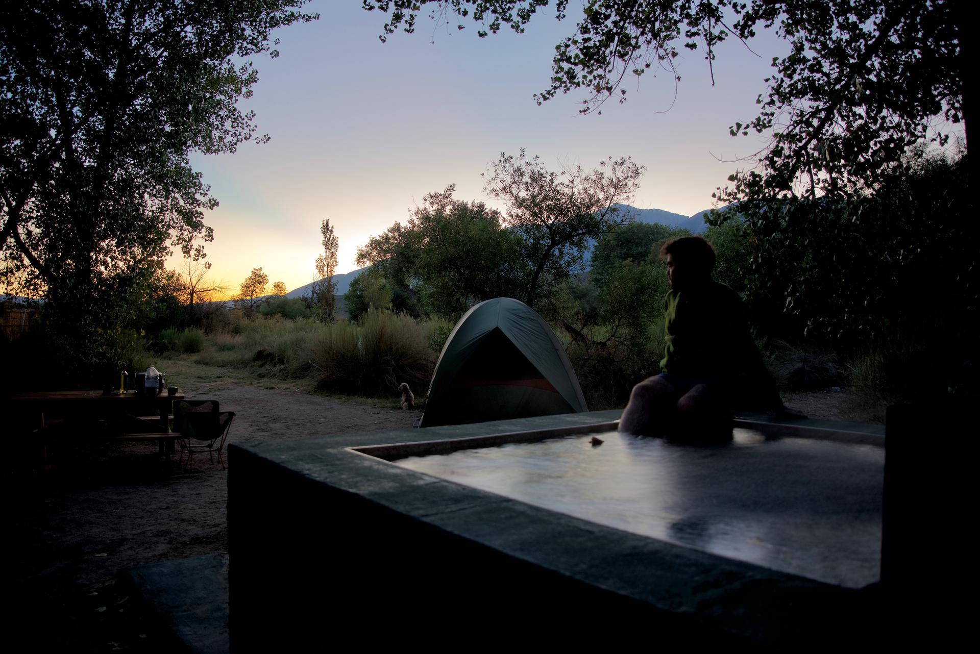 Person sits by a tent at dusk, with mountains in the distance.