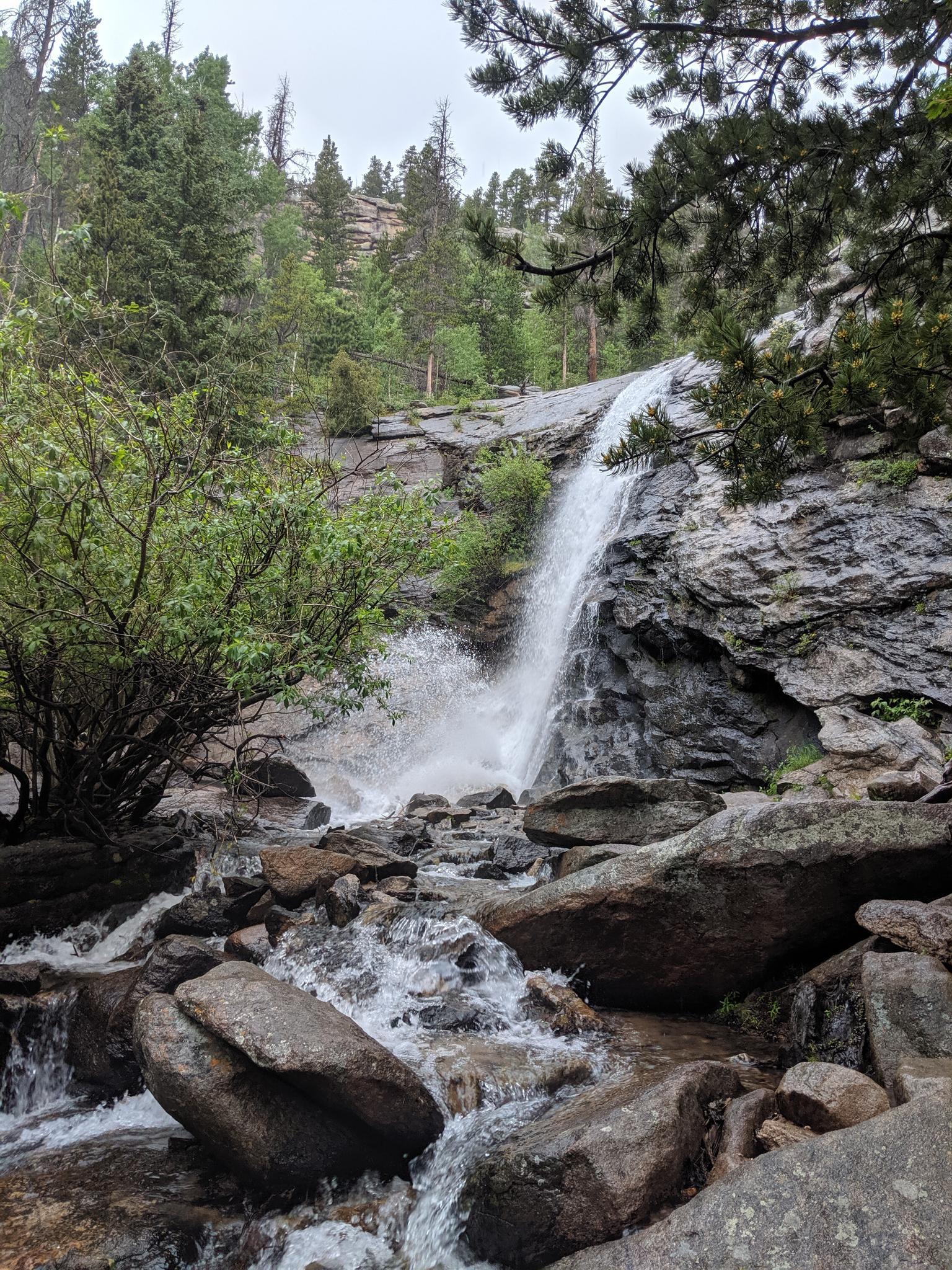 A powerful waterfall cascades over rocks into a stream under an overcast sky.