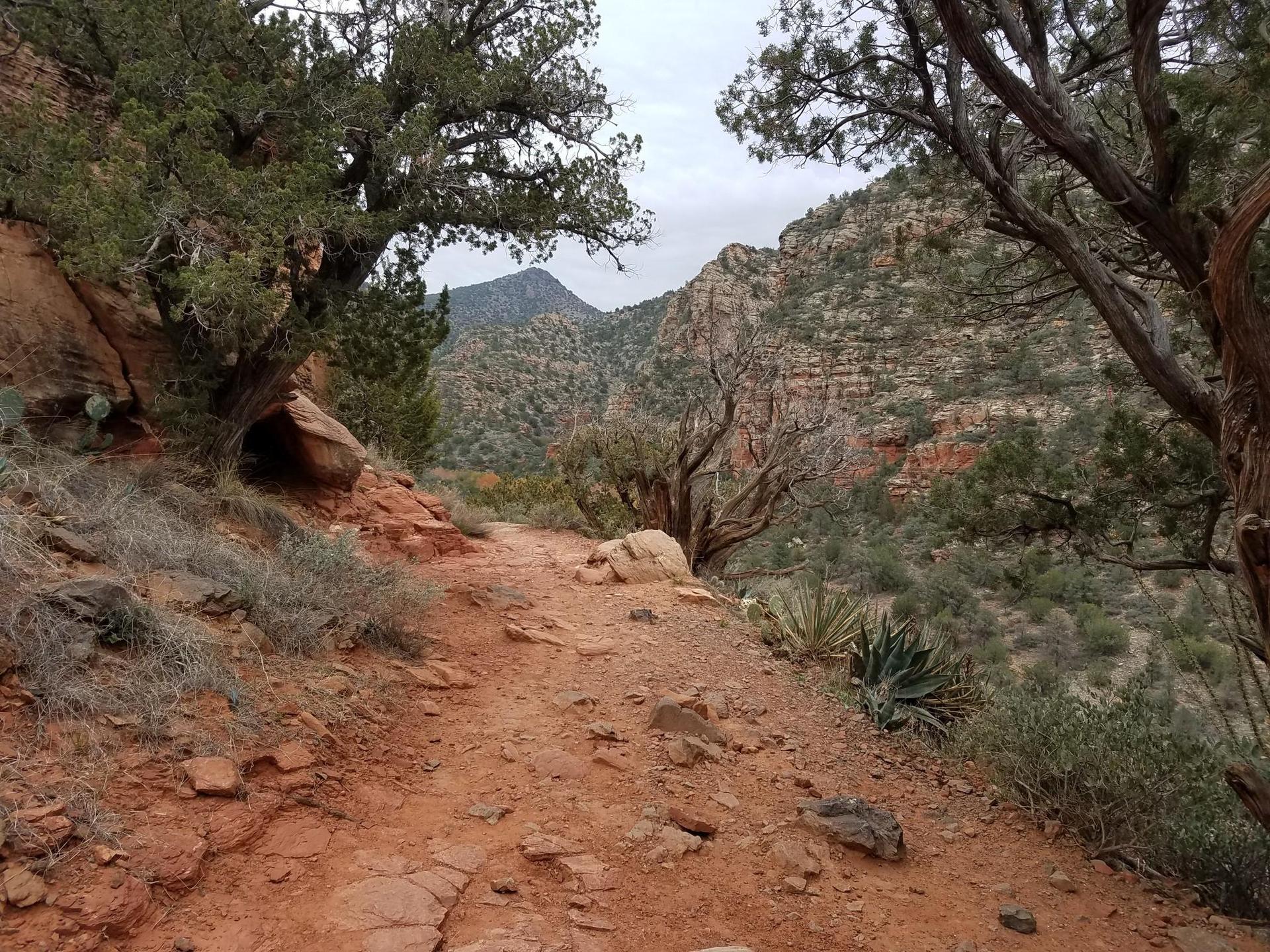 A winding red dirt path through rocky, arid mountains under an overcast sky.