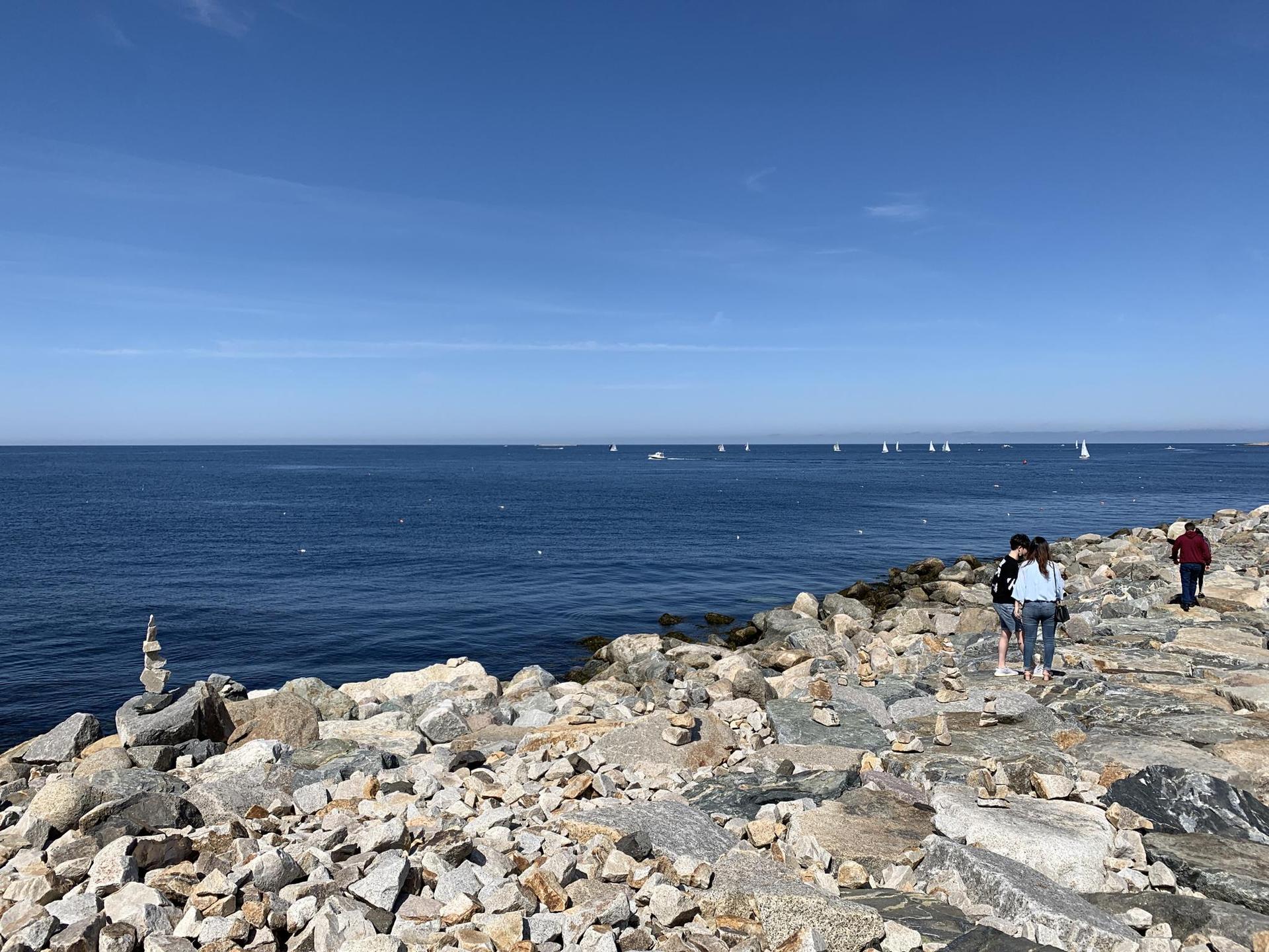People walk on a rocky shore beside a vast blue ocean with sailboats.