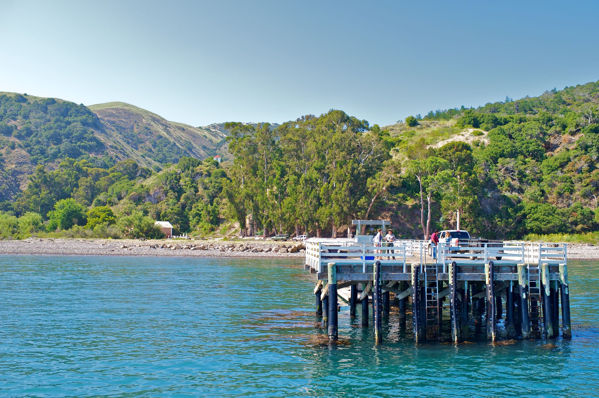Sunny view of a wooden pier with people, blue water, and green hills. - Del Norte Trail