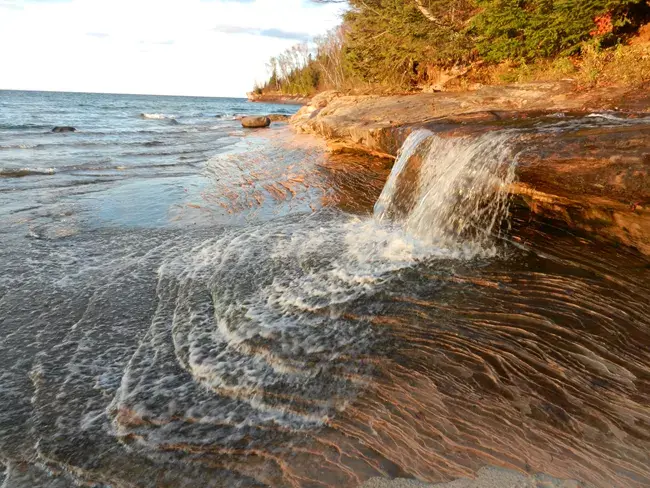 Small waterfall cascades over layered rock into a lake at golden hour.