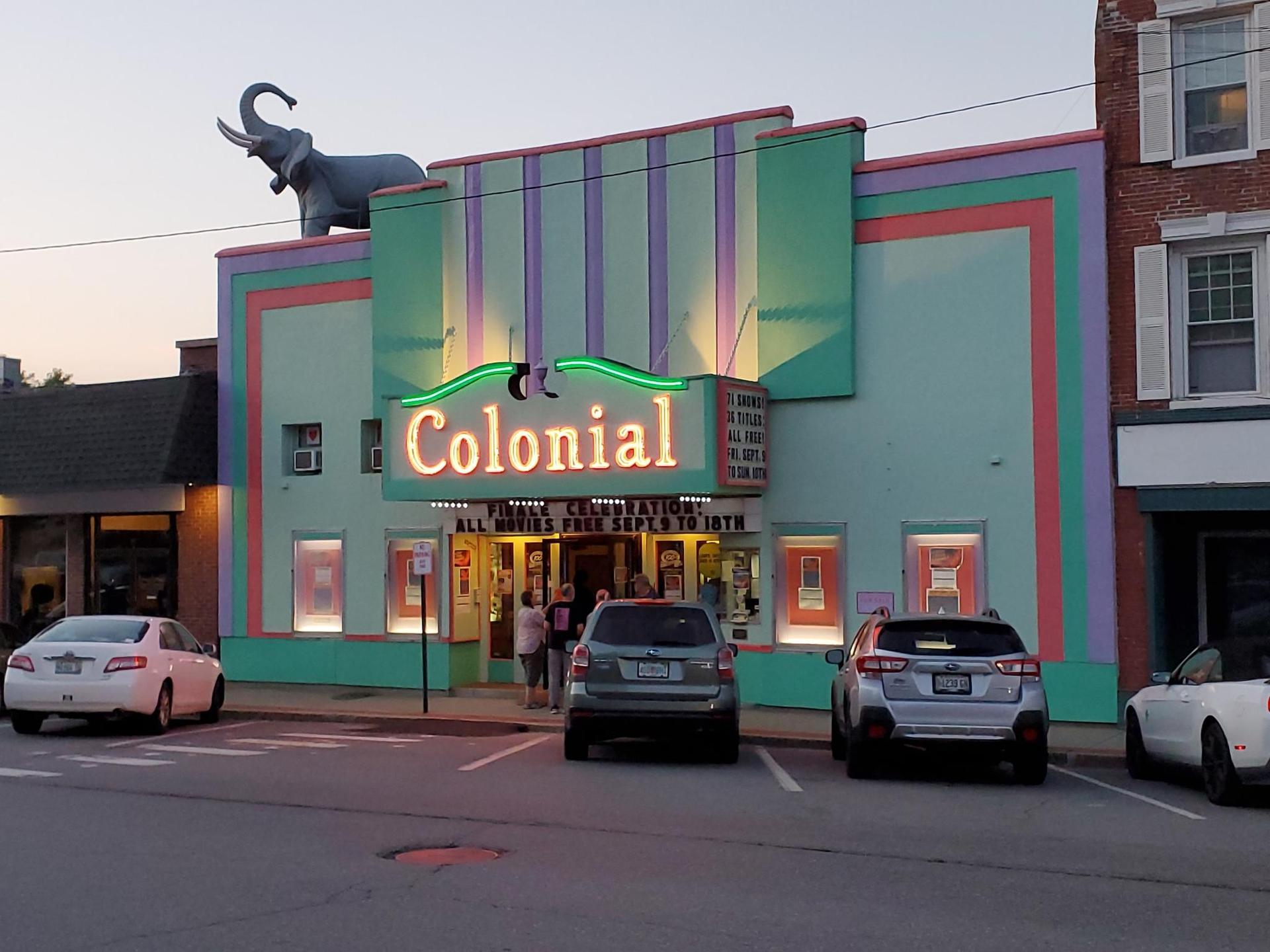 Colorful movie theater with elephant statue, neon lights, and people entering at dusk.