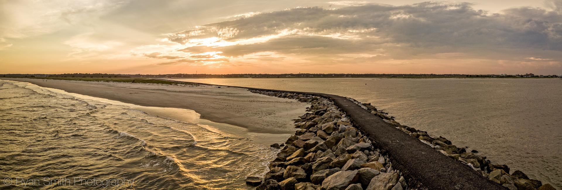 A long rock jetty separates ocean waves and calm water at sunset.