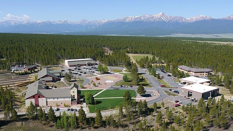 Aerial view of buildings nestled in a pine forest, with distant snow-capped mountains under bright sun.