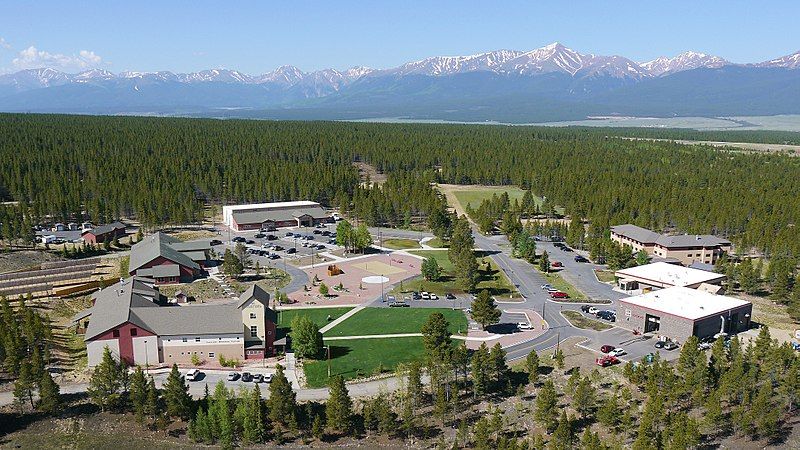 Aerial view of buildings nestled in a pine forest, with distant snow-capped mountains under bright sun. - Wheelers Way Loop