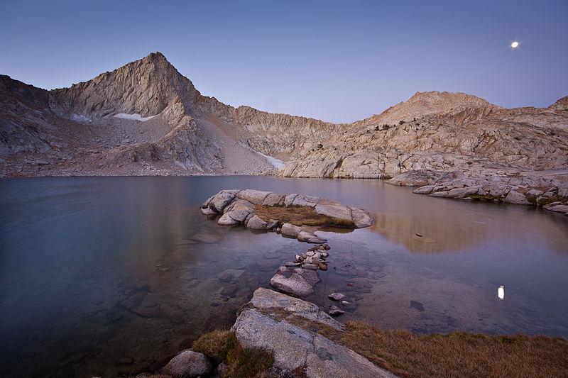 A serene mountain lake with rocky islands under a twilight sky with the moon.