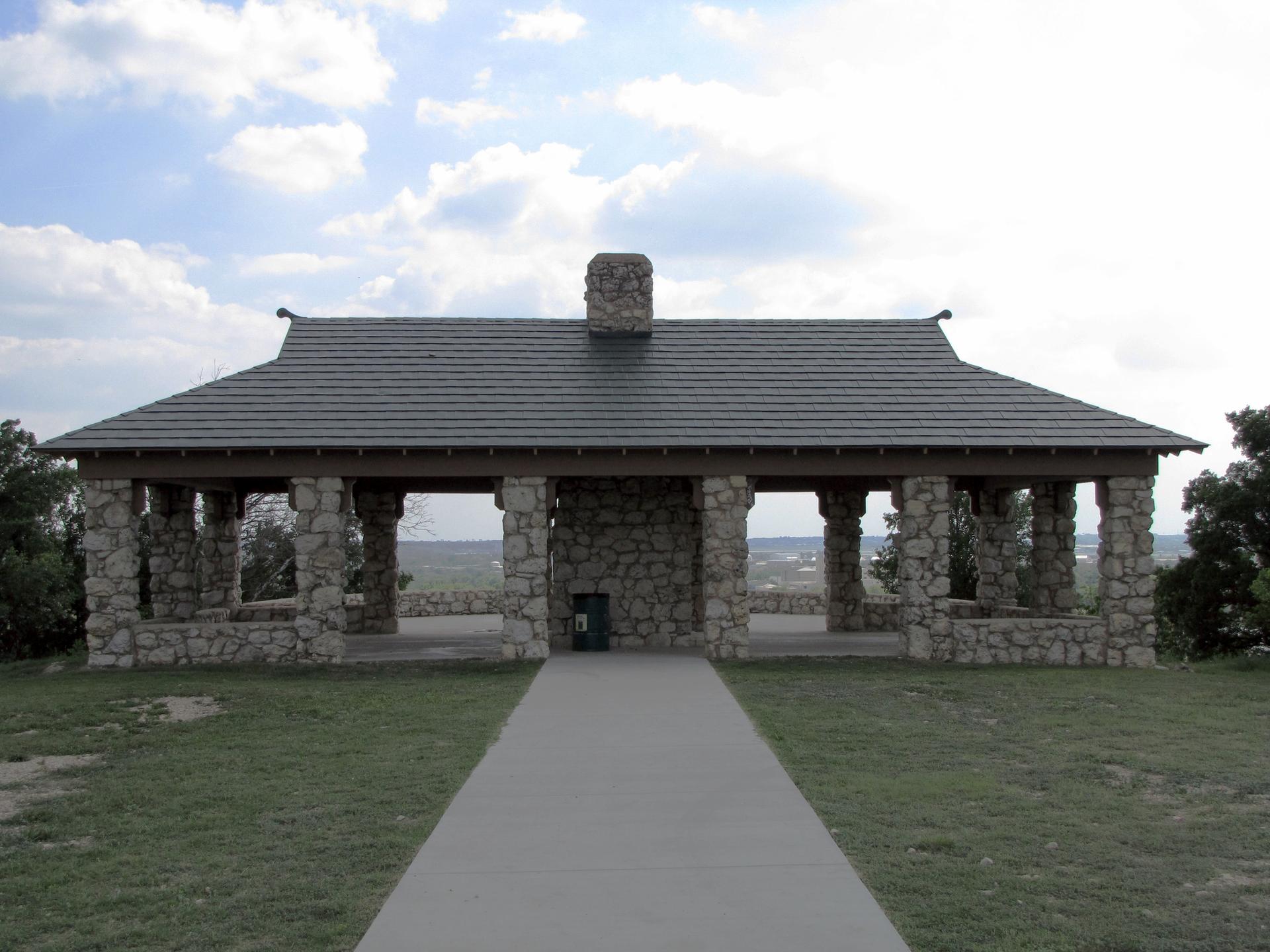 A large stone pavilion with a dark roof stands on a grassy hill under clouds.