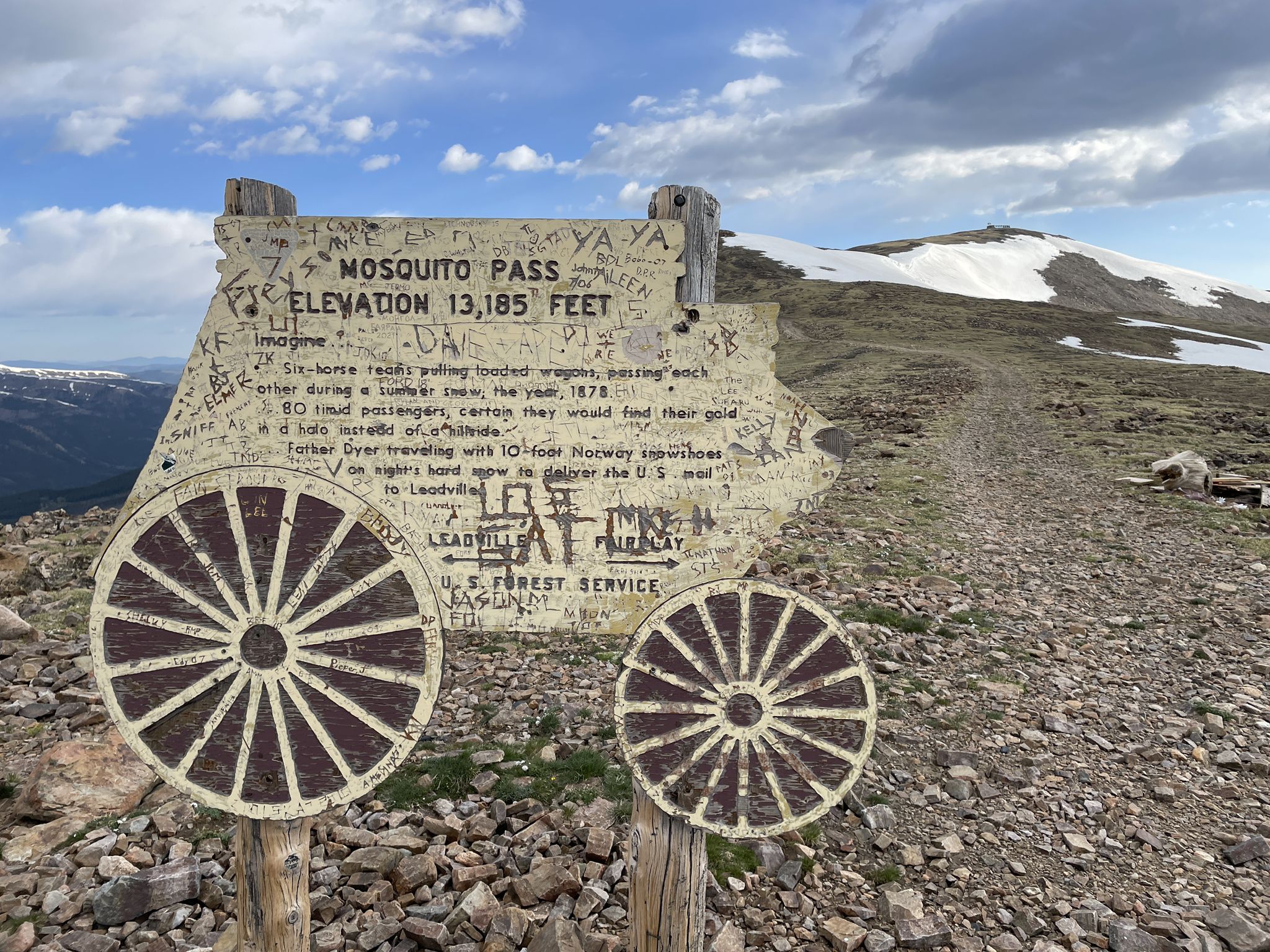 Weathered wooden sign with wagon wheels on a rocky mountain pass, snow-capped peak. - Mosquito Pass Road