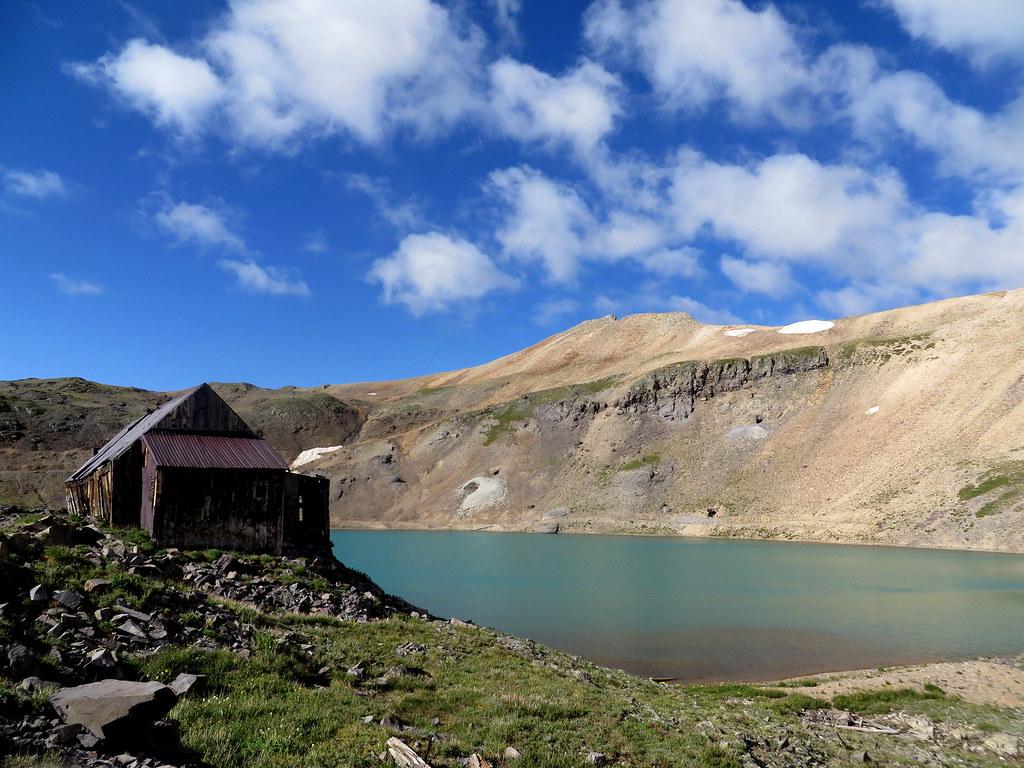Old building beside a bright blue lake with mountains under a sunny sky.