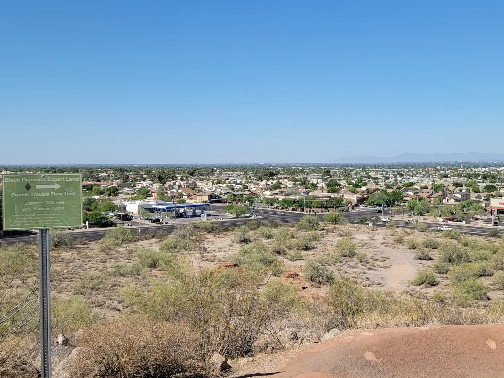 A trail sign overlooks a vast, sunny urban landscape with arid vegetation.