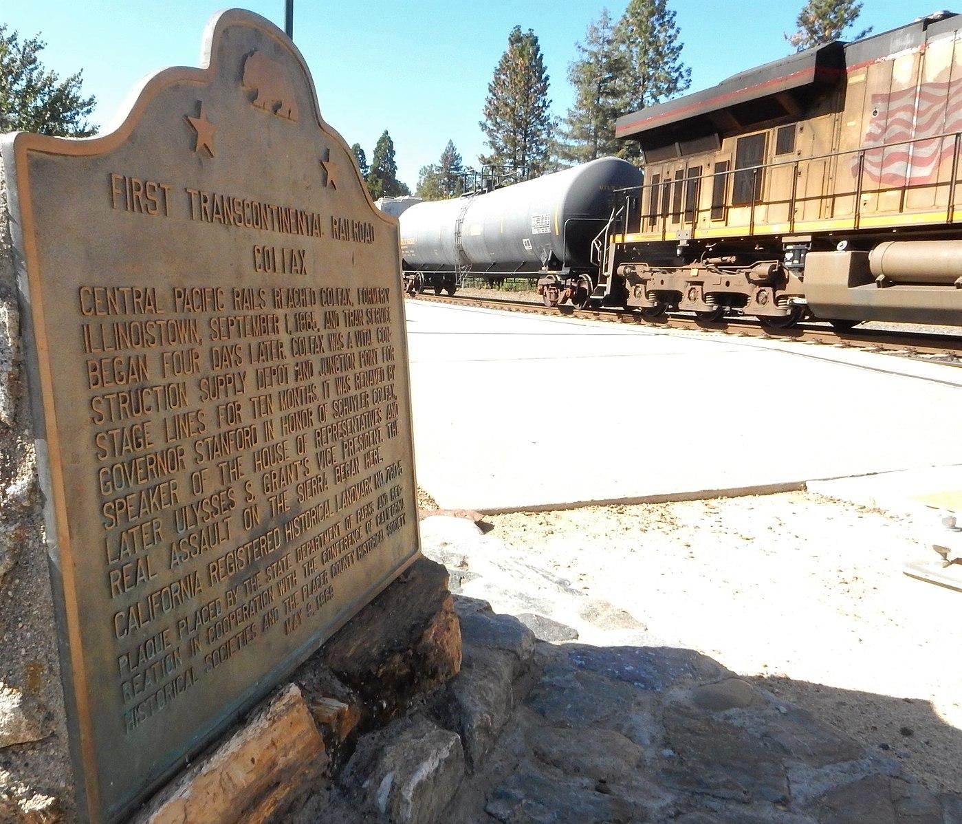 A bronze historical plaque in bright sunlight, with train cars behind it.